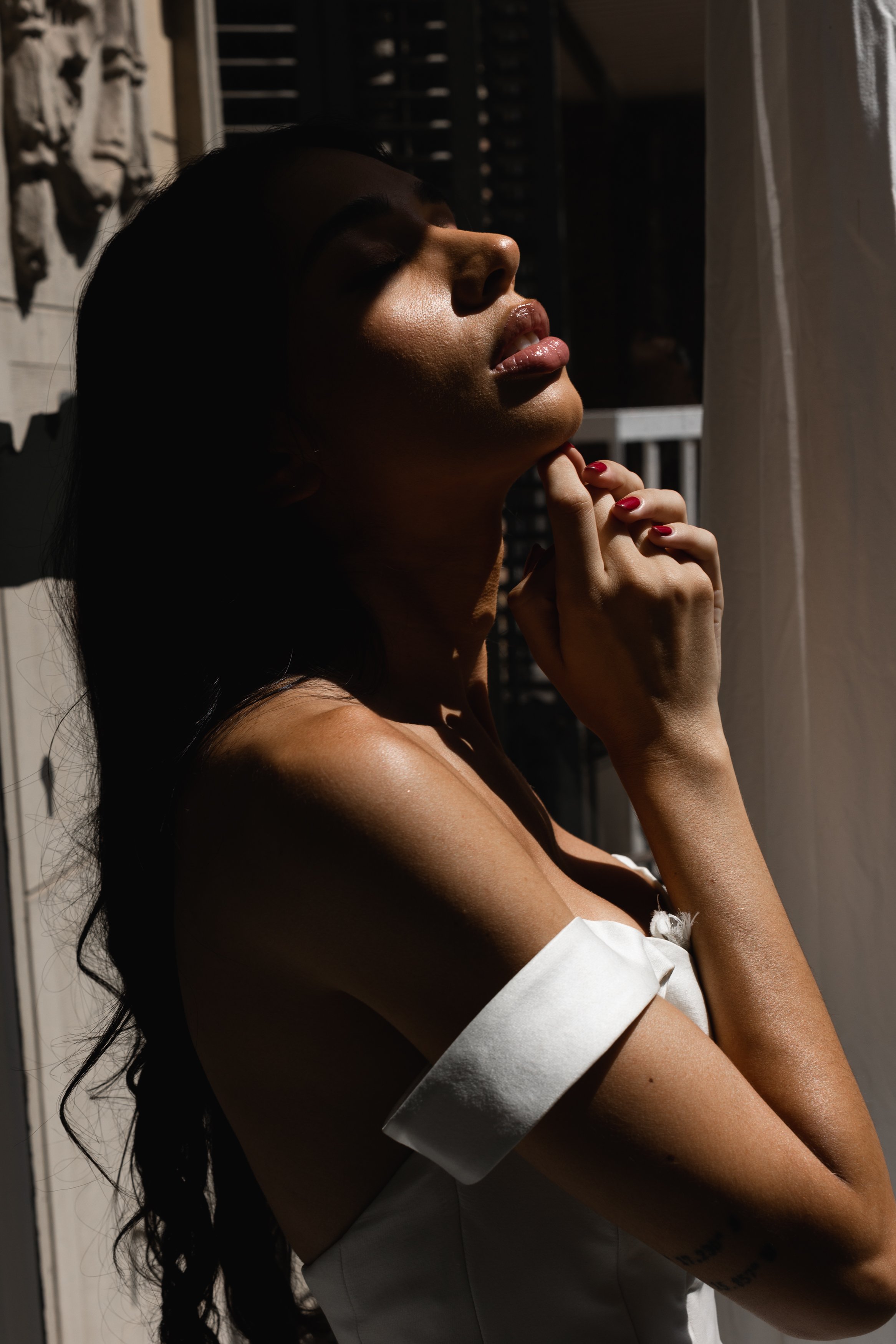 Side profile of a woman with dark hair, closed eyes, and glossy lips, standing near a window with sunlight creating shadows on her face.
