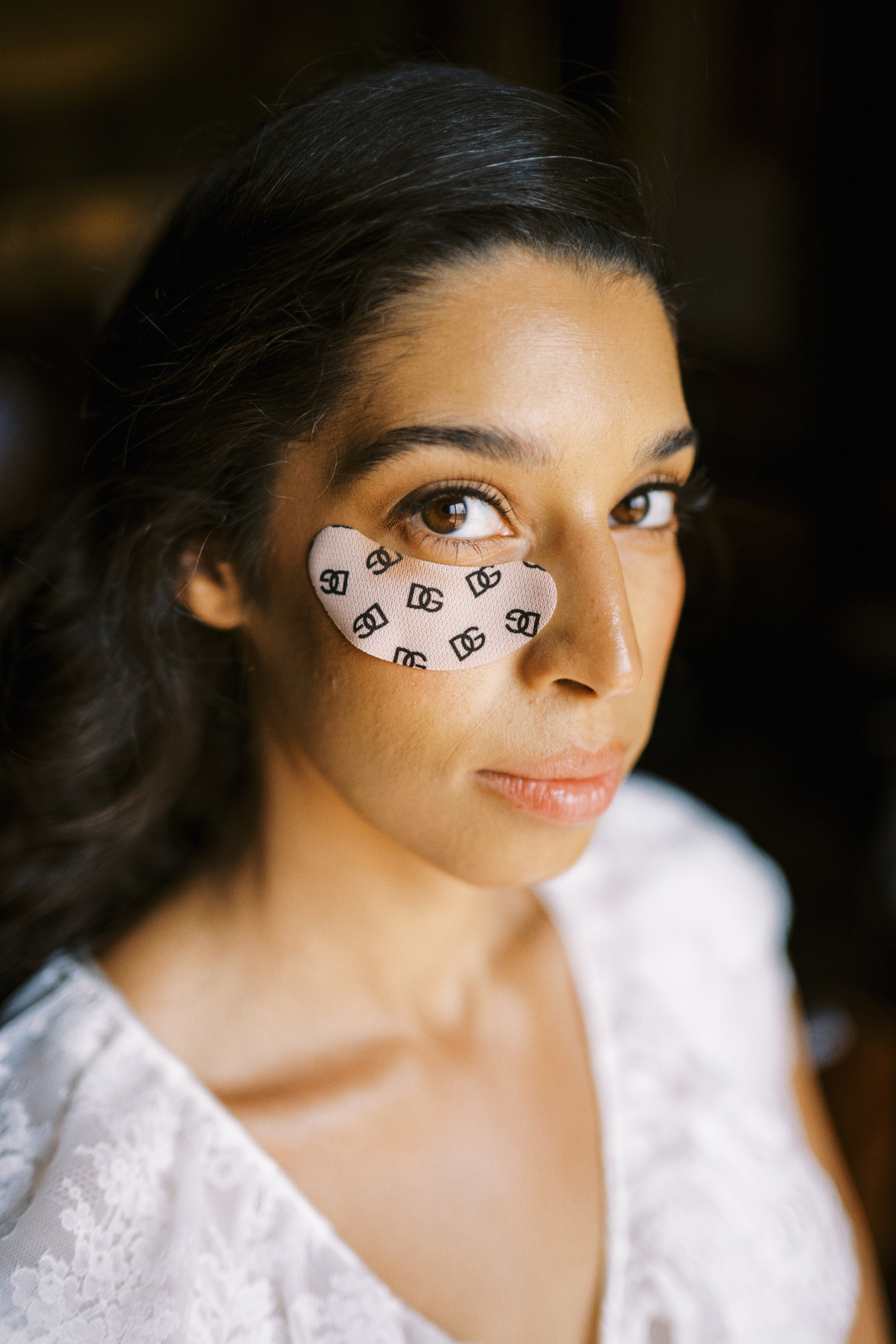 Close-up of a young woman with dark hair and brown eyes, wearing a heart-shaped patch with black logo pattern under her eye, dressed in a white lace top.