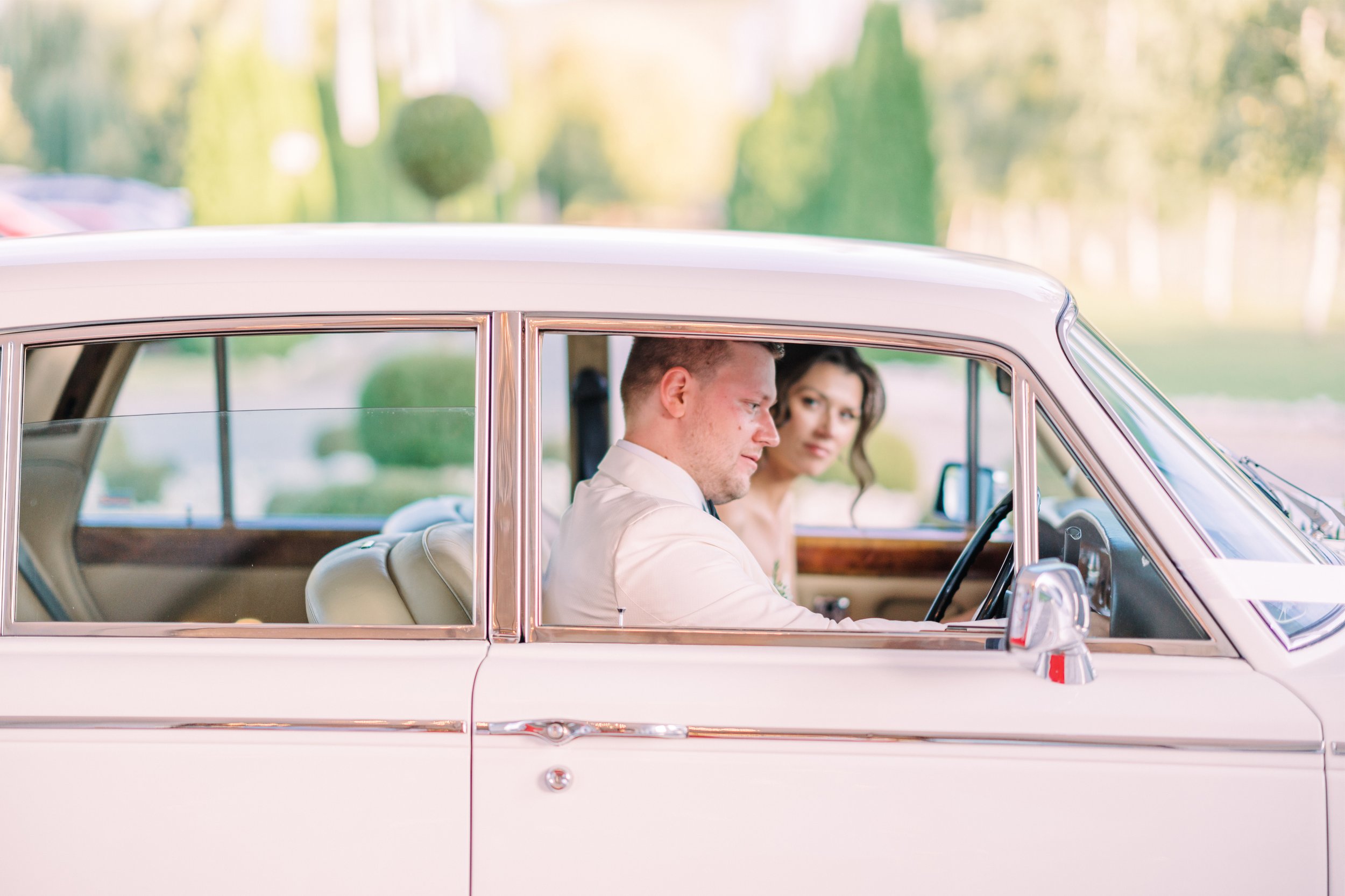 A man and woman sit inside a vintage white car, with the man driving and the woman looking at him, on a sunny day with blurred greenery in the background.