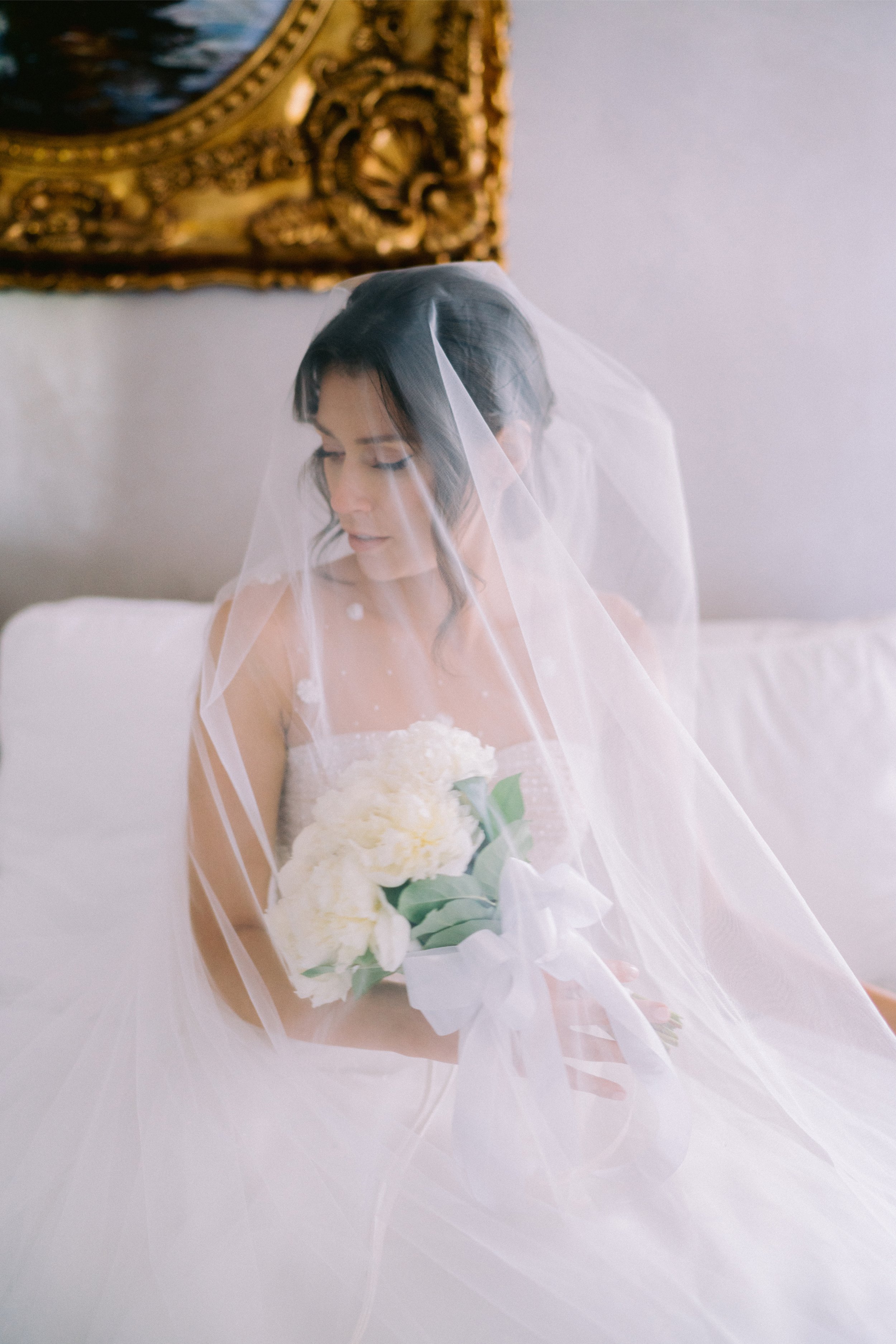 A bride with dark hair and a white wedding dress, wearing a veil, holding a bouquet of white flowers with green leaves, sitting on a white bed in a room with a gold-framed mirror on the wall.