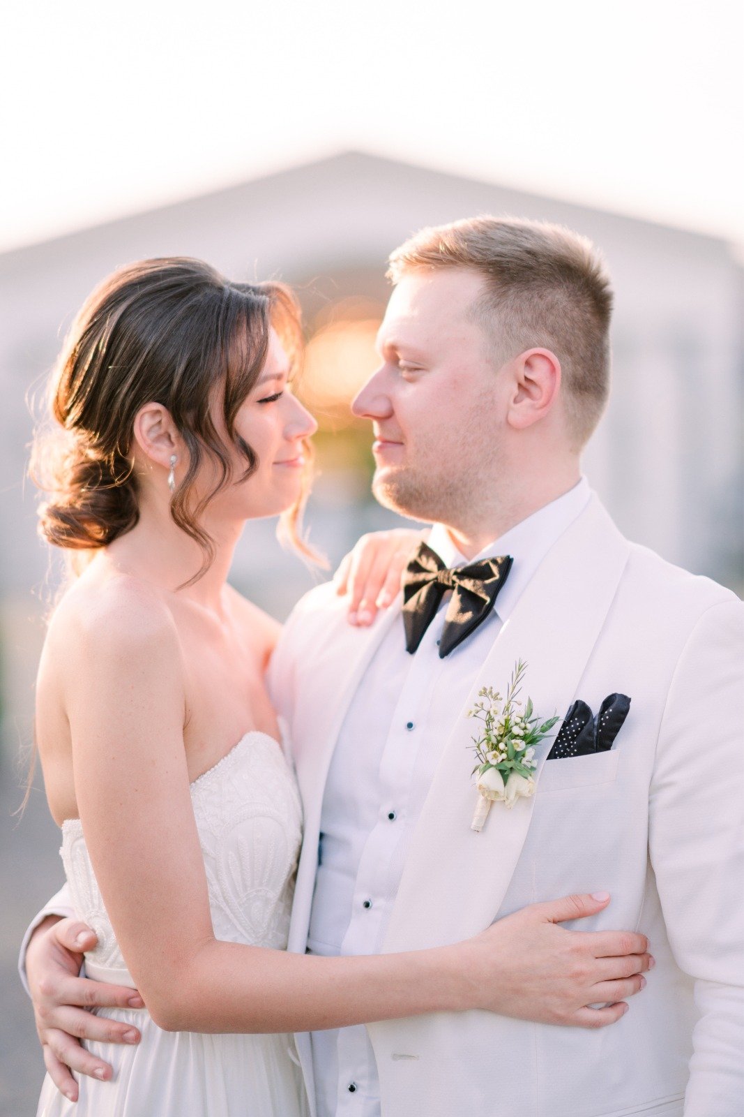 A bride and groom standing close, looking into each other's eyes, outdoors during sunset, with the bride wearing a strapless white wedding dress and the groom in a white tuxedo with a black bow tie.
