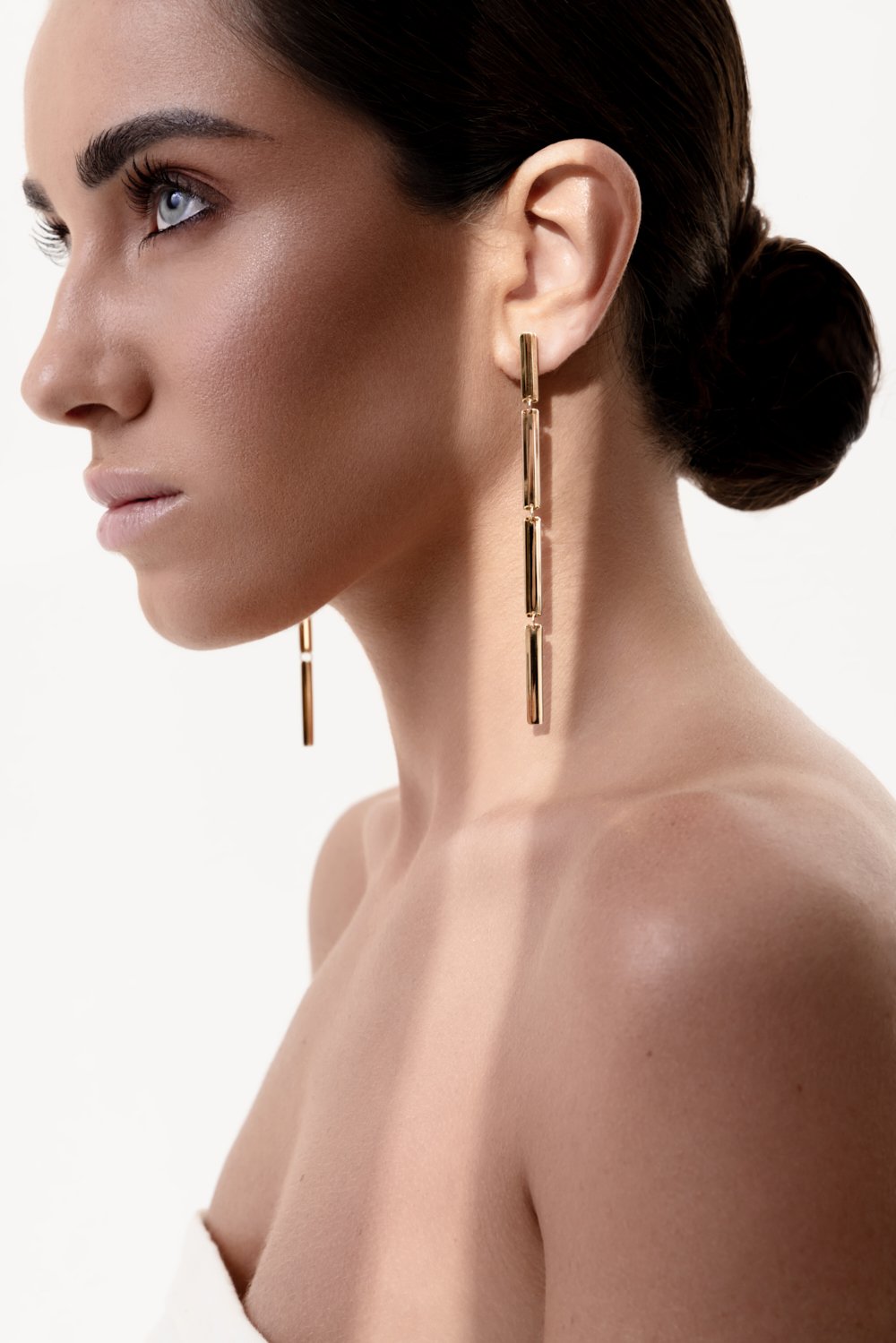 Close-up of a woman with dark hair in a bun, wearing large dangling gold earrings and minimal makeup against a plain white background.