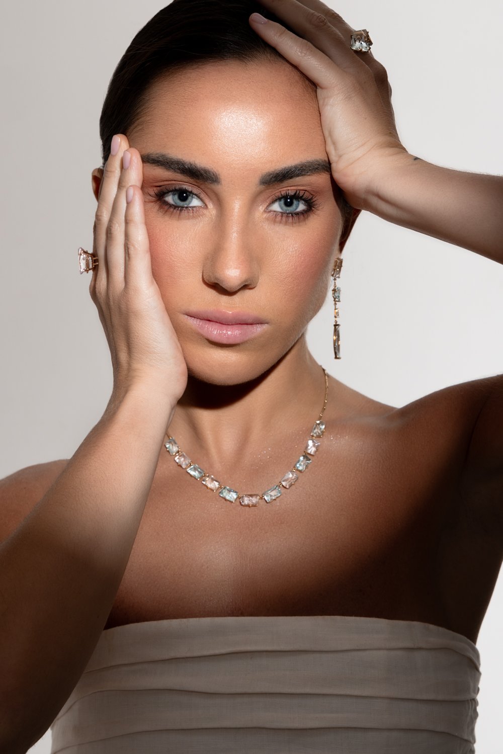 A woman with blue eyes, dark hair, and bare shoulders wearing diamond jewelry, including a necklace, earrings, and rings, posing with her hands on her head and face.