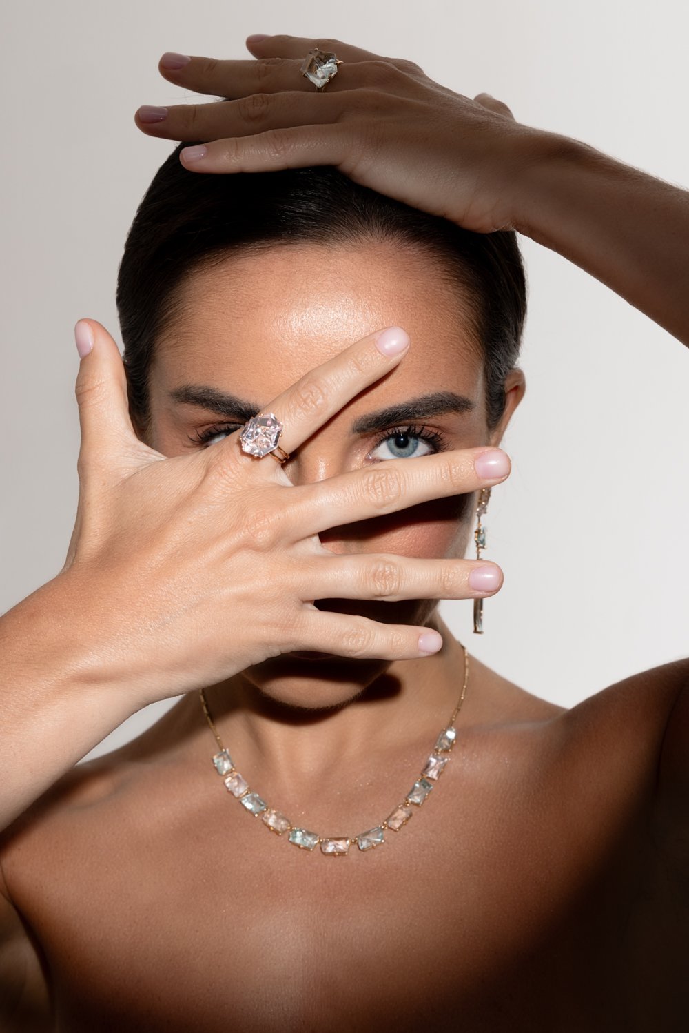A woman with blue eyes and dark hair, wearing a sparkle necklace and a large ring, covers part of her face with her hand, highlighting her eye and jewelry.