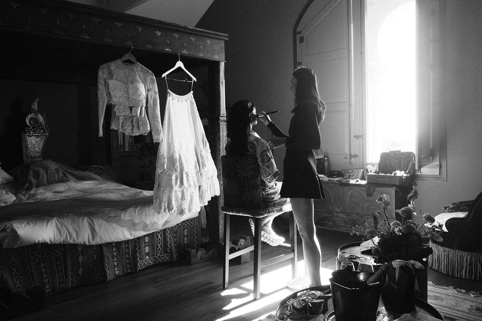 A woman sitting on a chair while another woman applies makeup to her face. There are hanging clothes, including a lace dress, on the wardrobe. The room is brightly lit by sunlight coming through open shutters. Flowers are visible on a table and a bed.