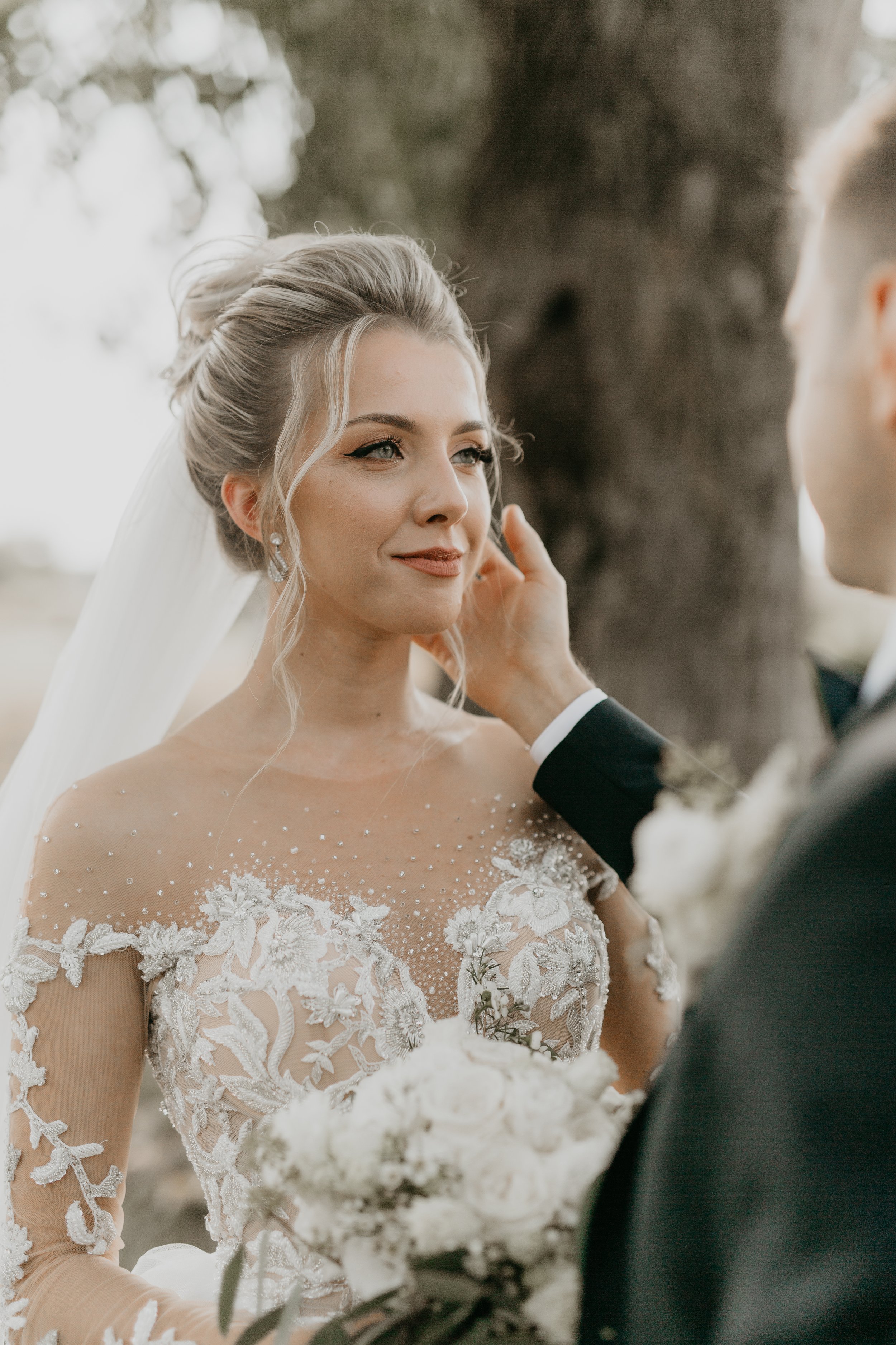 A bride with blonde hair styled in an updo and wearing a lace wedding dress with floral embroidery is looking at her groom with a gentle smile. She is wearing pearl earrings and a veil. The groom, partially visible, is wearing a black tuxedo and is t