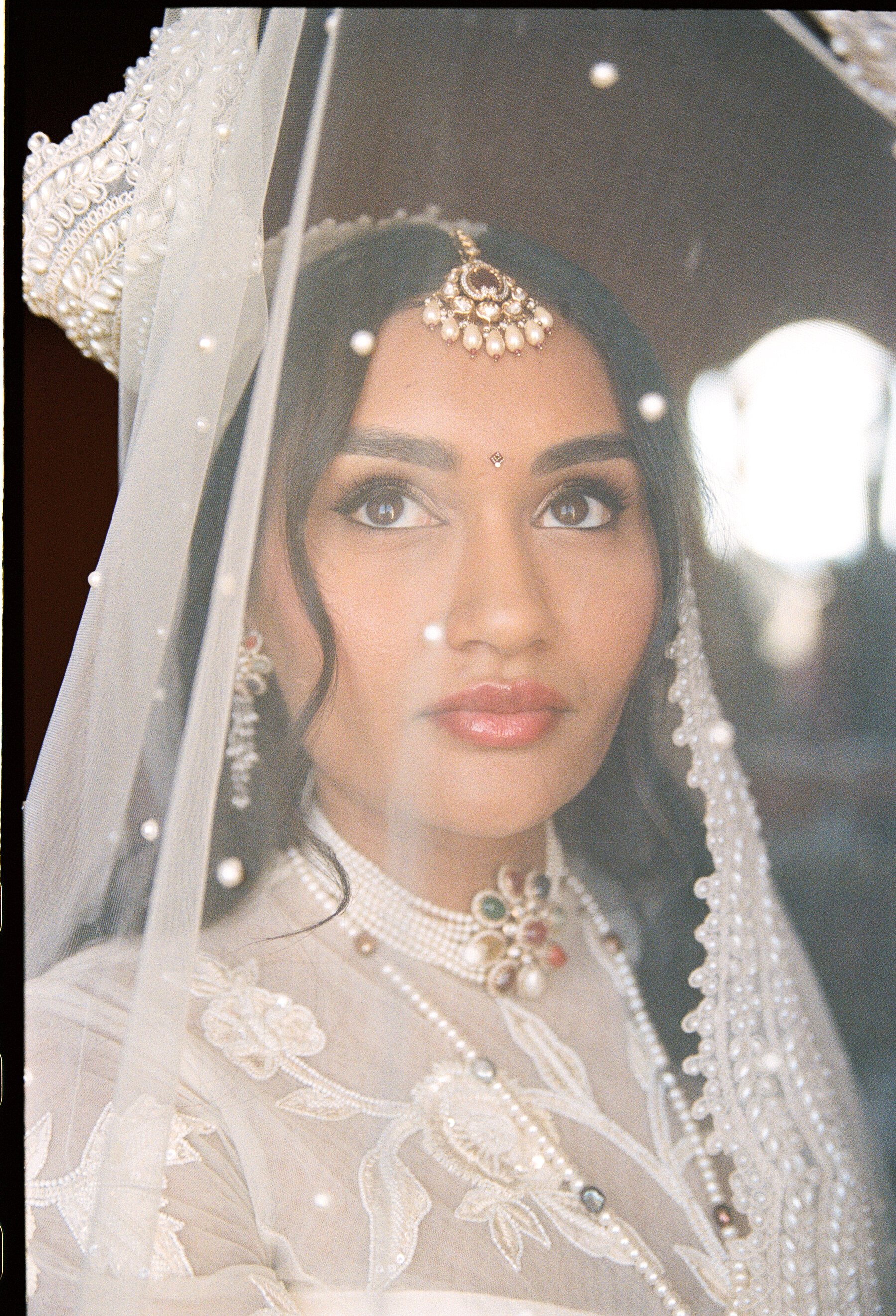 A woman dressed in white bride attire, wearing traditional jewelry including a headpiece and necklace, seen through a bridal veil with pearl and embellishment details.