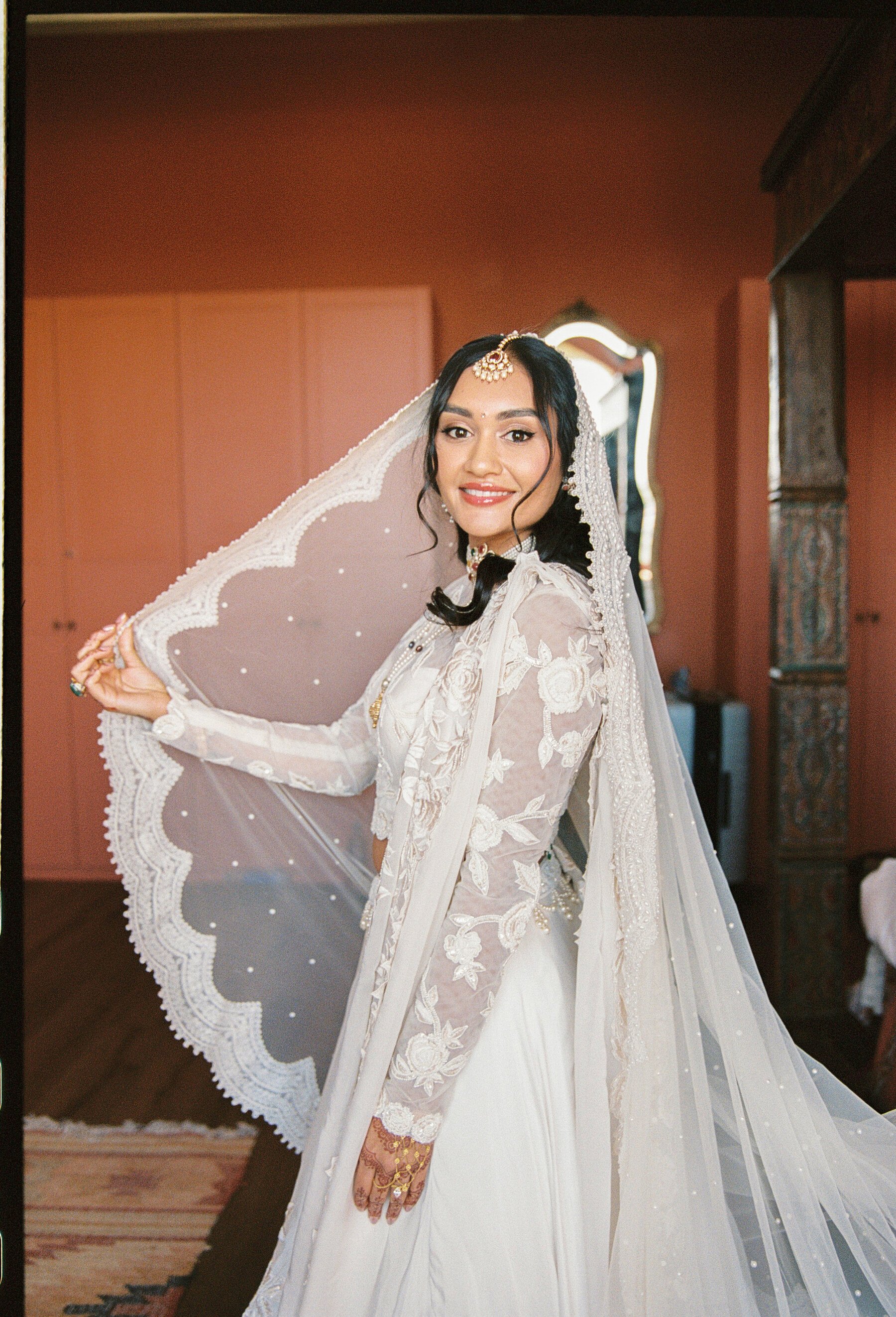 A bride in a white wedding dress and traditional jewelry is holding a lace parasol and smiling. She has dark hair styled with soft curls and a veil, with a wooden column and ornate mirror in the background.