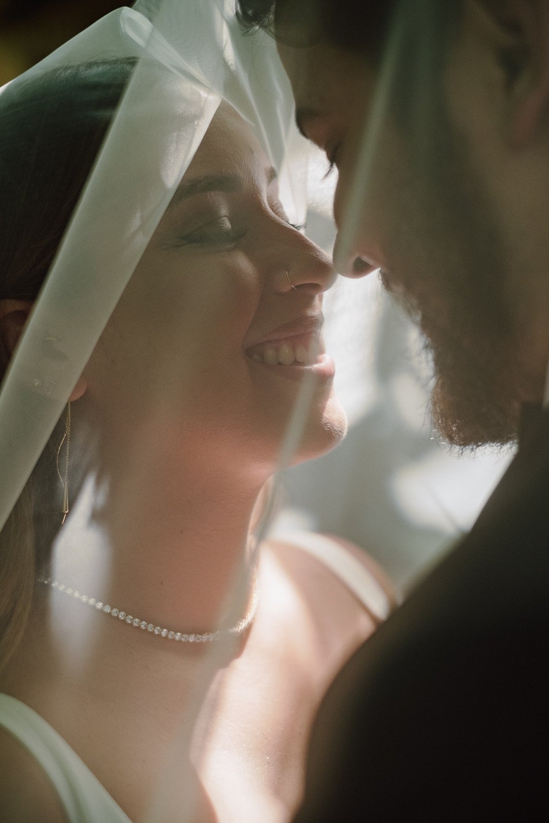 Close-up of a couple about to kiss, with the woman smiling and wearing a veil, and the man with a beard, both with their faces close.
