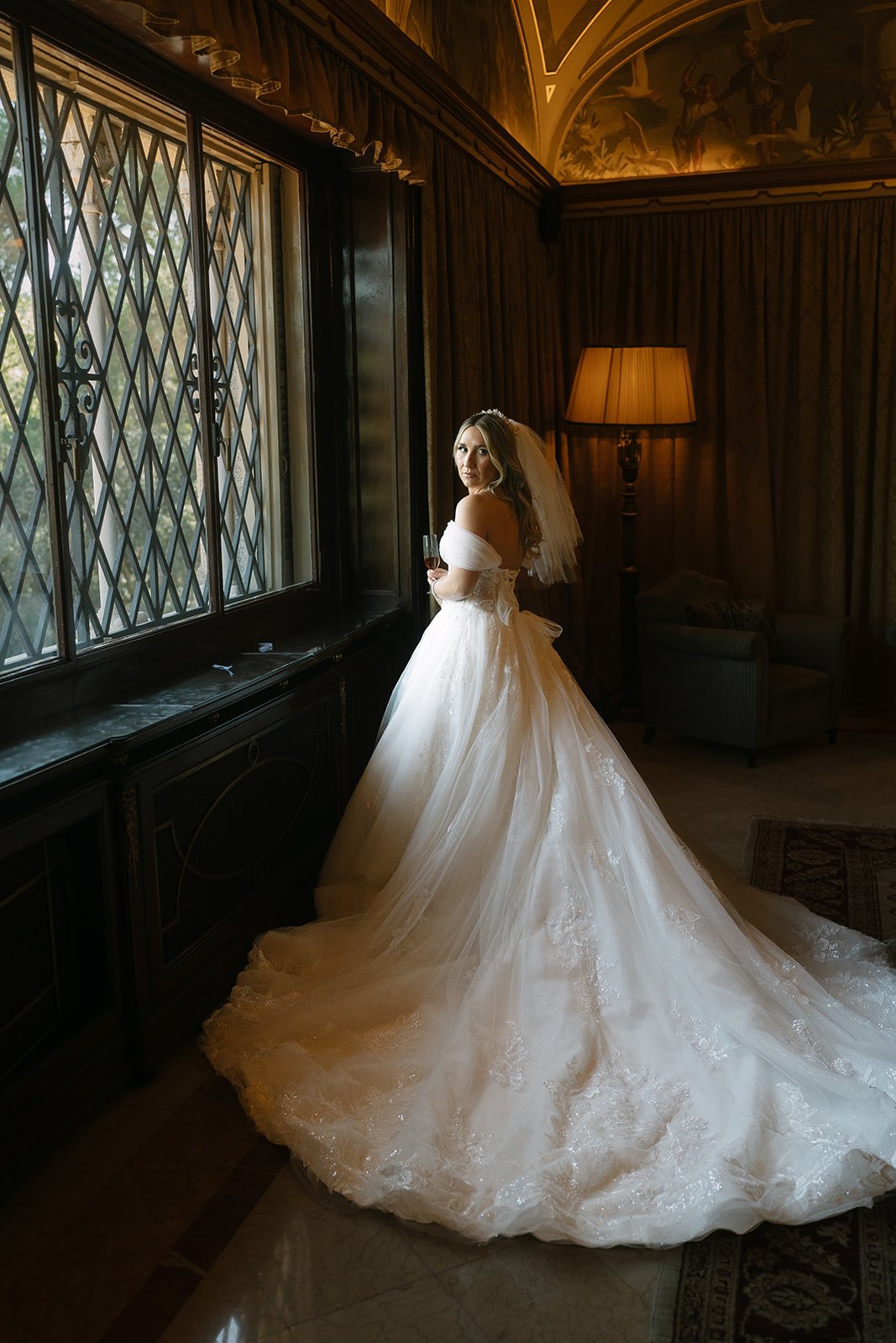 A bride in a white wedding gown with off-the-shoulder sleeves, holding a glass, standing near a window with decorative ironwork, in a richly decorated room with dark wood paneling, curtains, a floor lamp, and a tapestry ceiling painting.