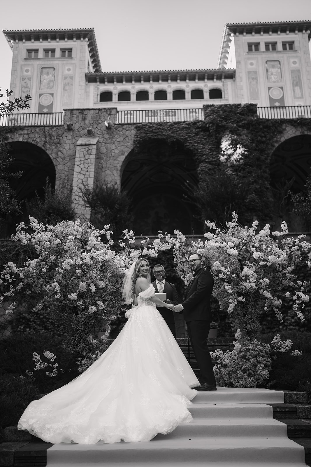 A black and white photo of a wedding ceremony outdoors, with a bride and groom holding hands and smiling, standing on stairs in front of flowering bushes, with a large ornate building in the background.