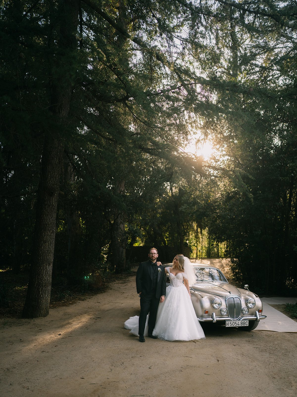 A bride and groom standing in front of a vintage silver car on a wooded path with sunlight filtering through the trees.
