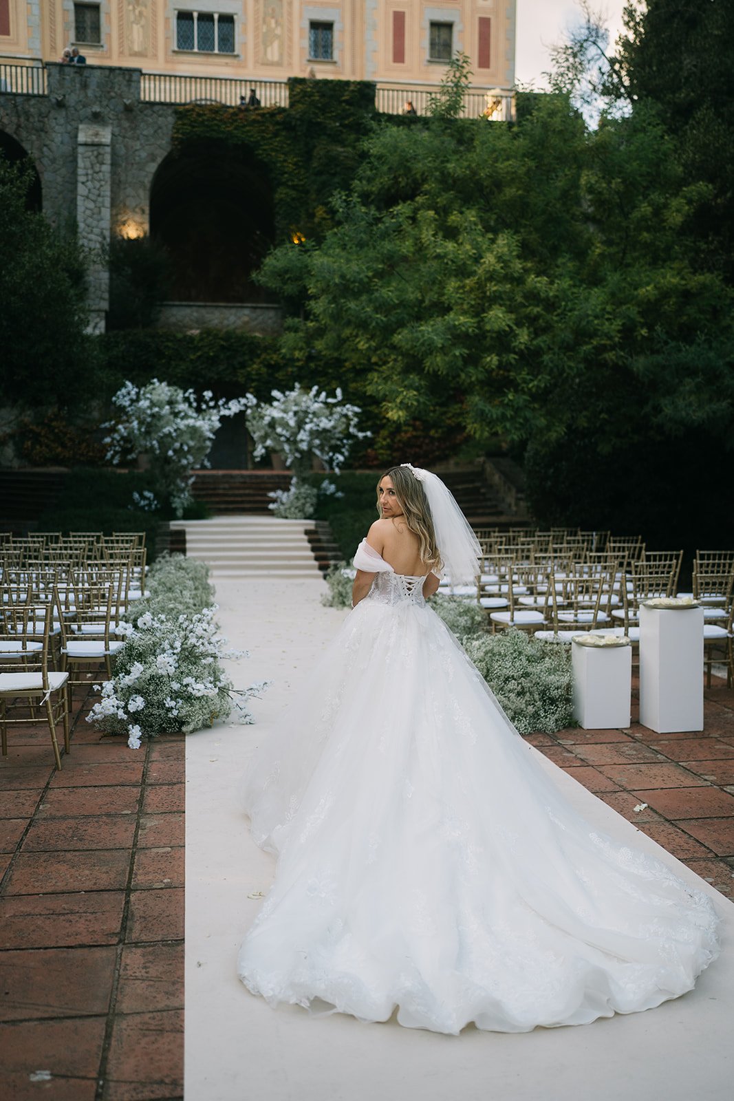 Bride in a wedding gown standing on an aisle decorated with white flowers, outdoors with trees and a building in the background.