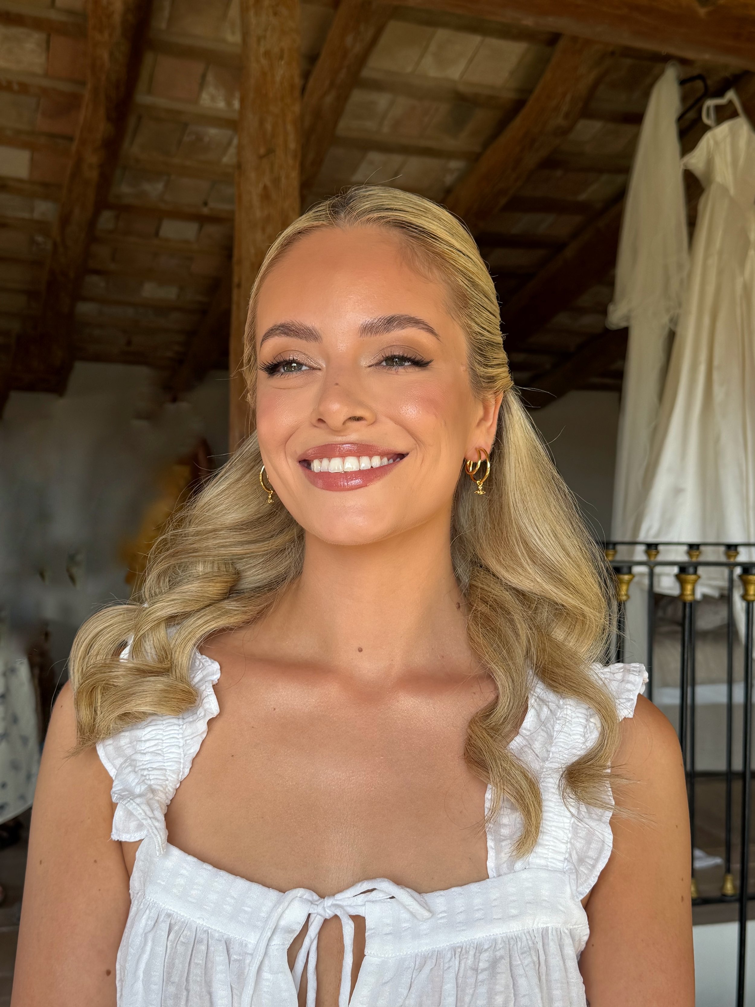 A smiling woman with long blonde hair and gold hoop earrings, wearing a white ruffled dress, indoors in a rustic setting with wooden beams and a partially visible white dress hanging in the background.