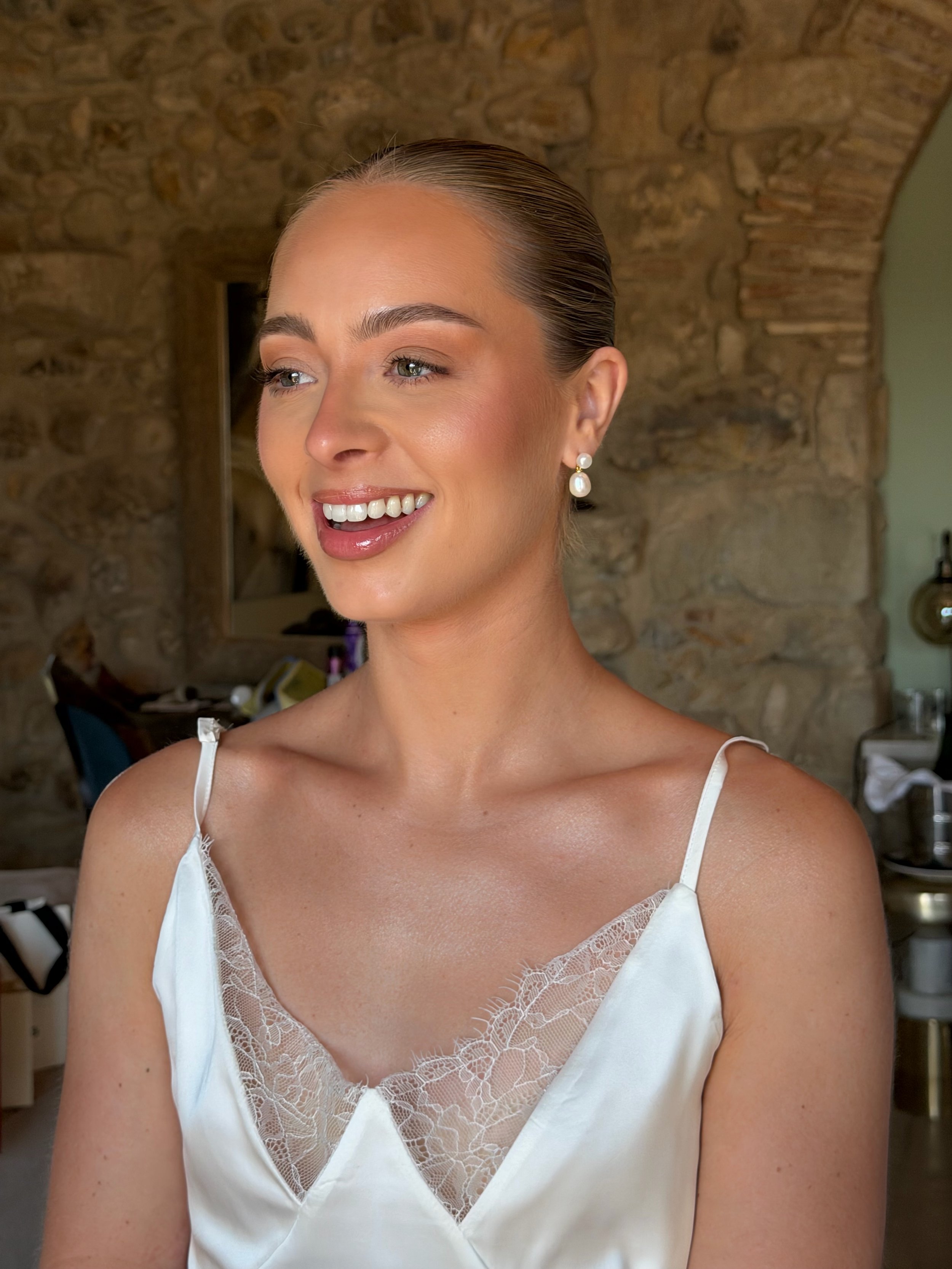 A smiling woman with styled hair, wearing pearl earrings and a white satin dress with lace details, no makeup makeup look, skin focused makeup,done by roberta boldea.