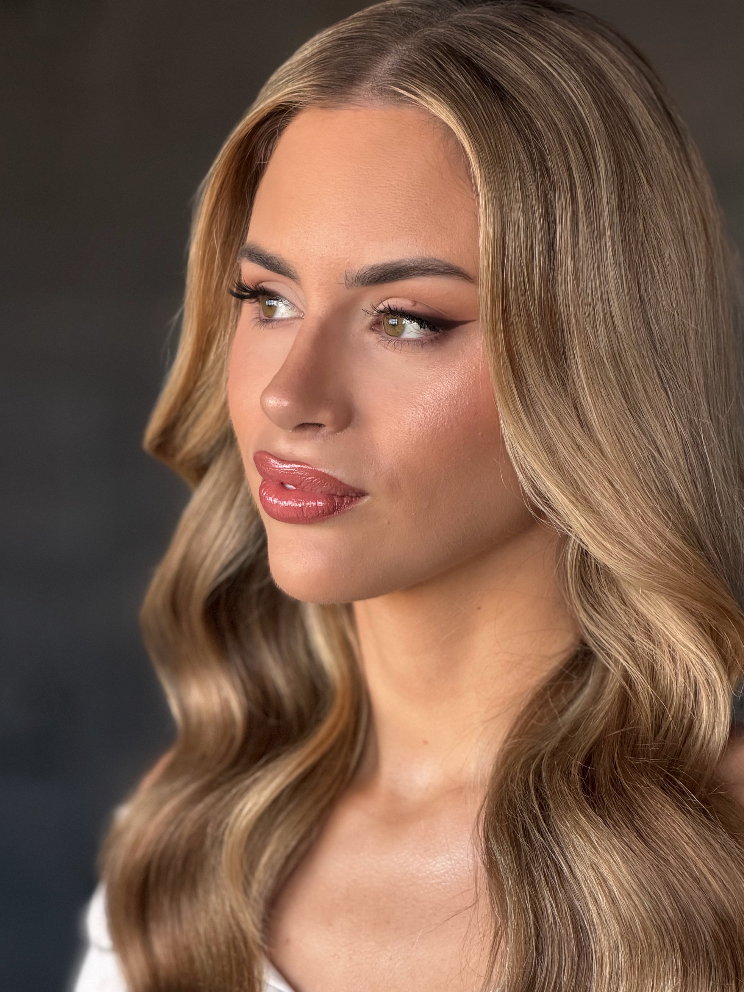 Close-up of a woman with long, wavy, light brown hair and subtle makeup, looking to the side. Soft Glam makeup, skin focused look