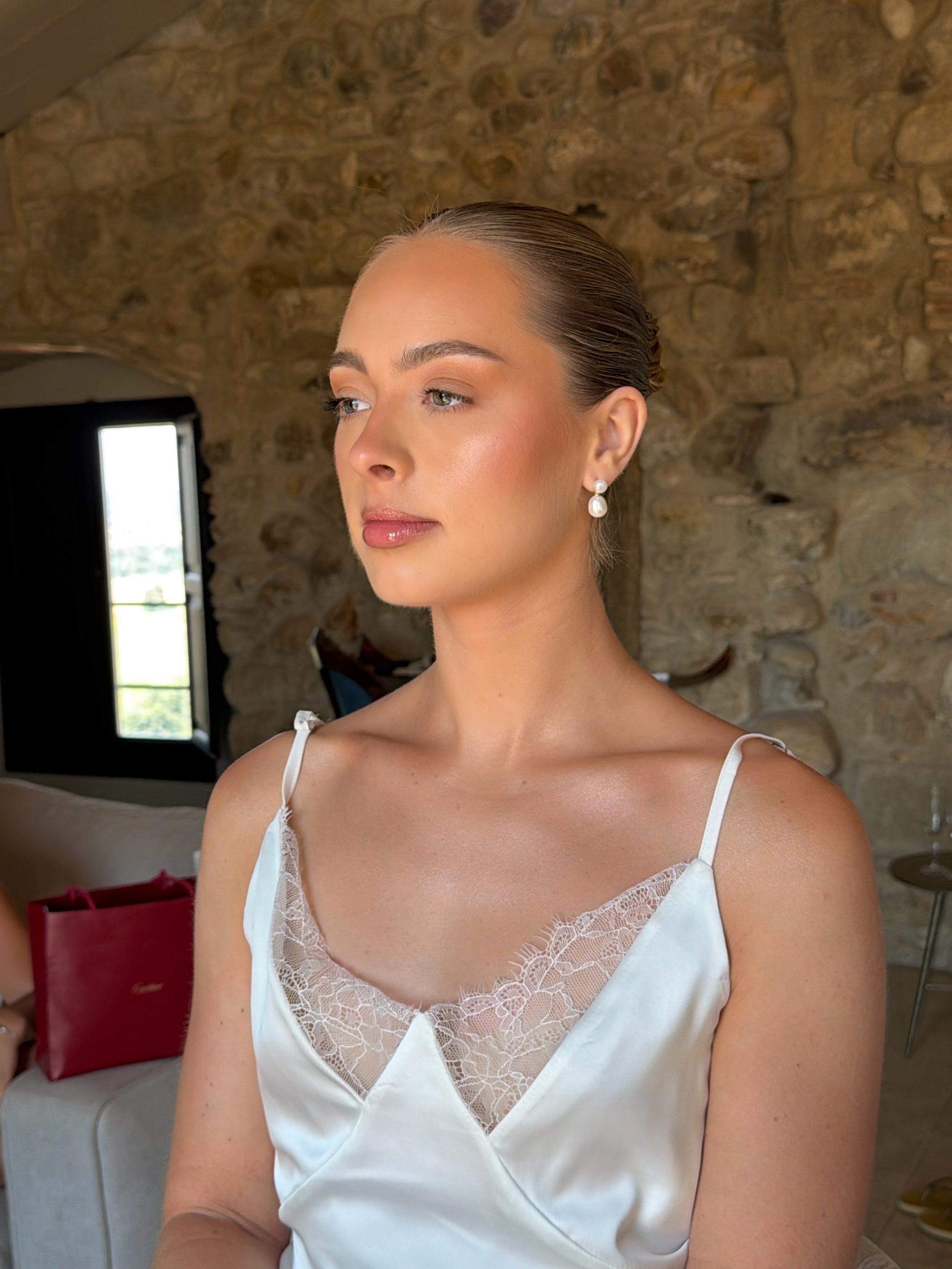 A woman with fair skin and light eyes, wearing pearl earrings and a white satin nightgown with lace trim, stands against a rustic stone wall interior. Her hair is slicked back, and she gazes to the side with a calm expression.