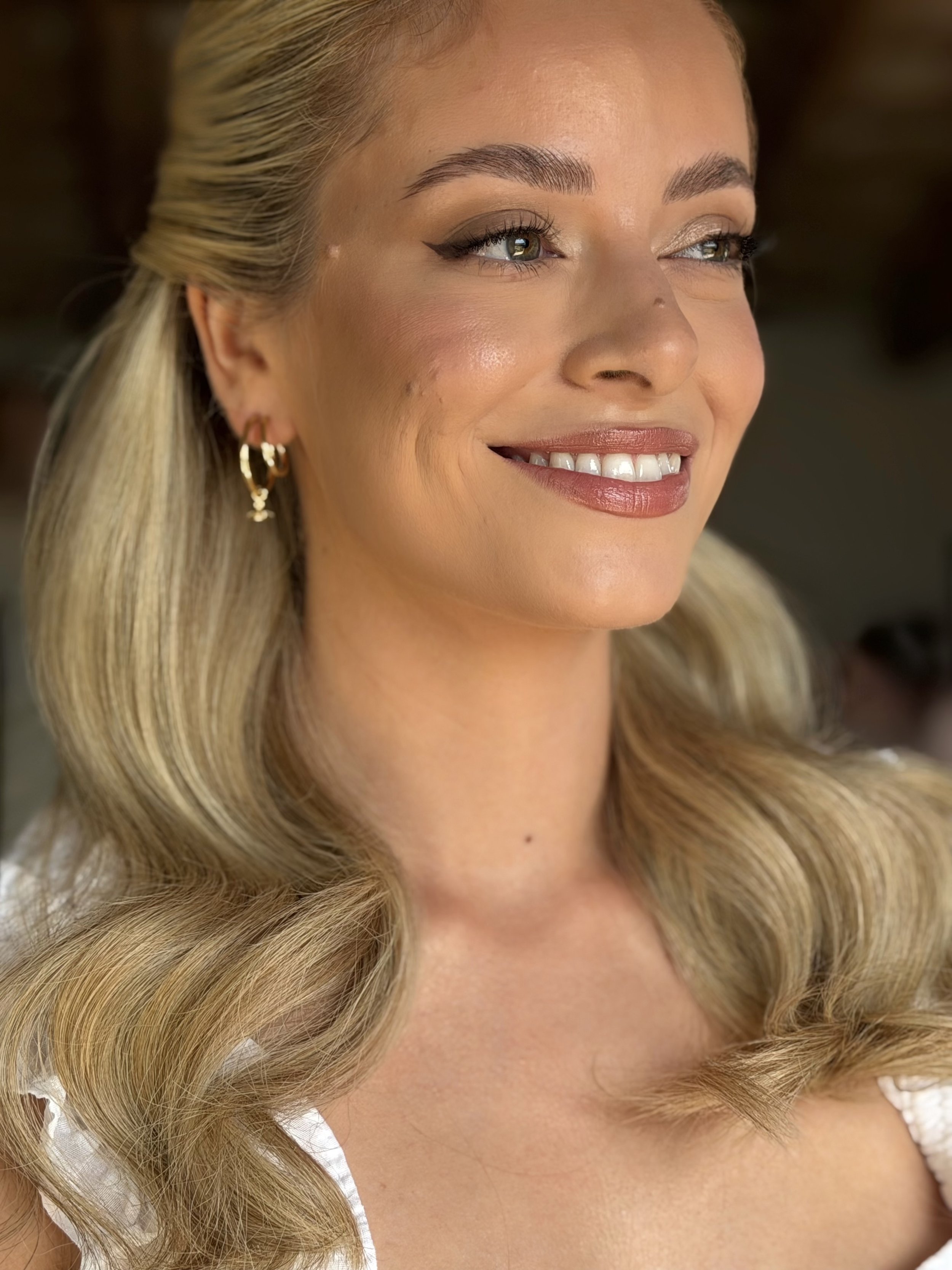 Close-up of a smiling woman with blonde wavy hair, light facemakeup, winged eyeliner, modern bridal look.