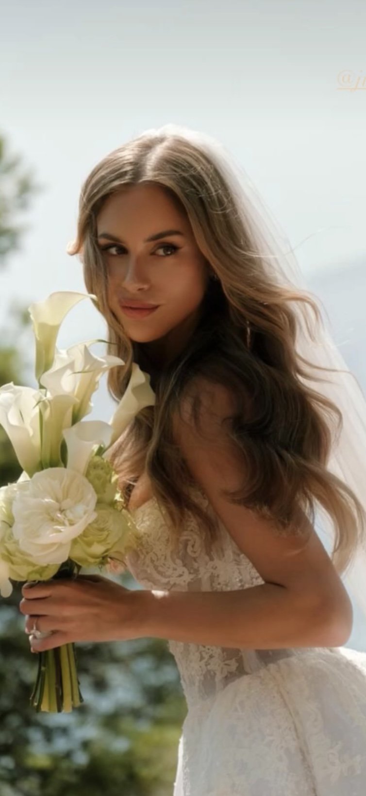 A woman in a wedding dress holding a bouquet of white flowers with a blurred outdoor background.