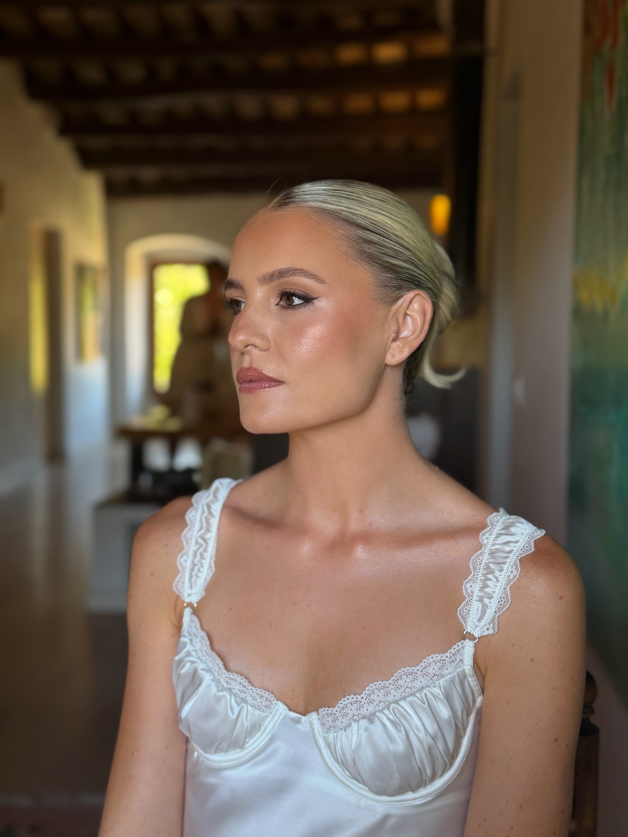 A woman with blonde hair slicked back and makeup, wearing a white satin top with lace straps, standing in a room with wooden ceiling beams and decorative wall art. Winged eyeliner 