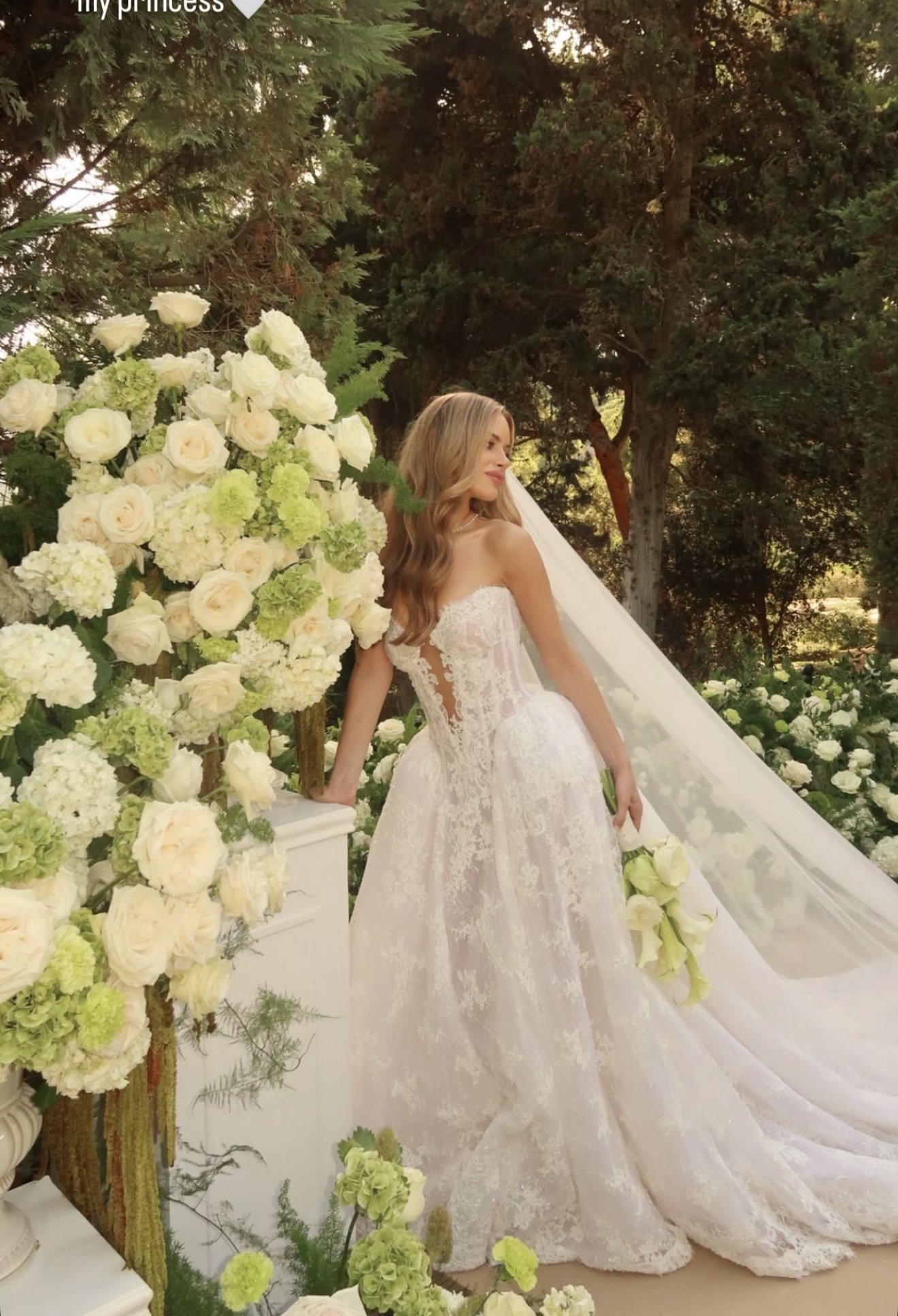 A bride in a white wedding dress with lace details, standing outdoors among greenery and white flowers, Bride in soft glam with neutral colors, medium skin tone, full covarage, modern bridal look