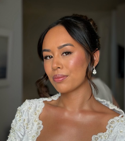 Close-up of a woman with dark hair styled in an updo, wearing earrings and a white dress with intricate lace detailing, looking confidently at the camera.