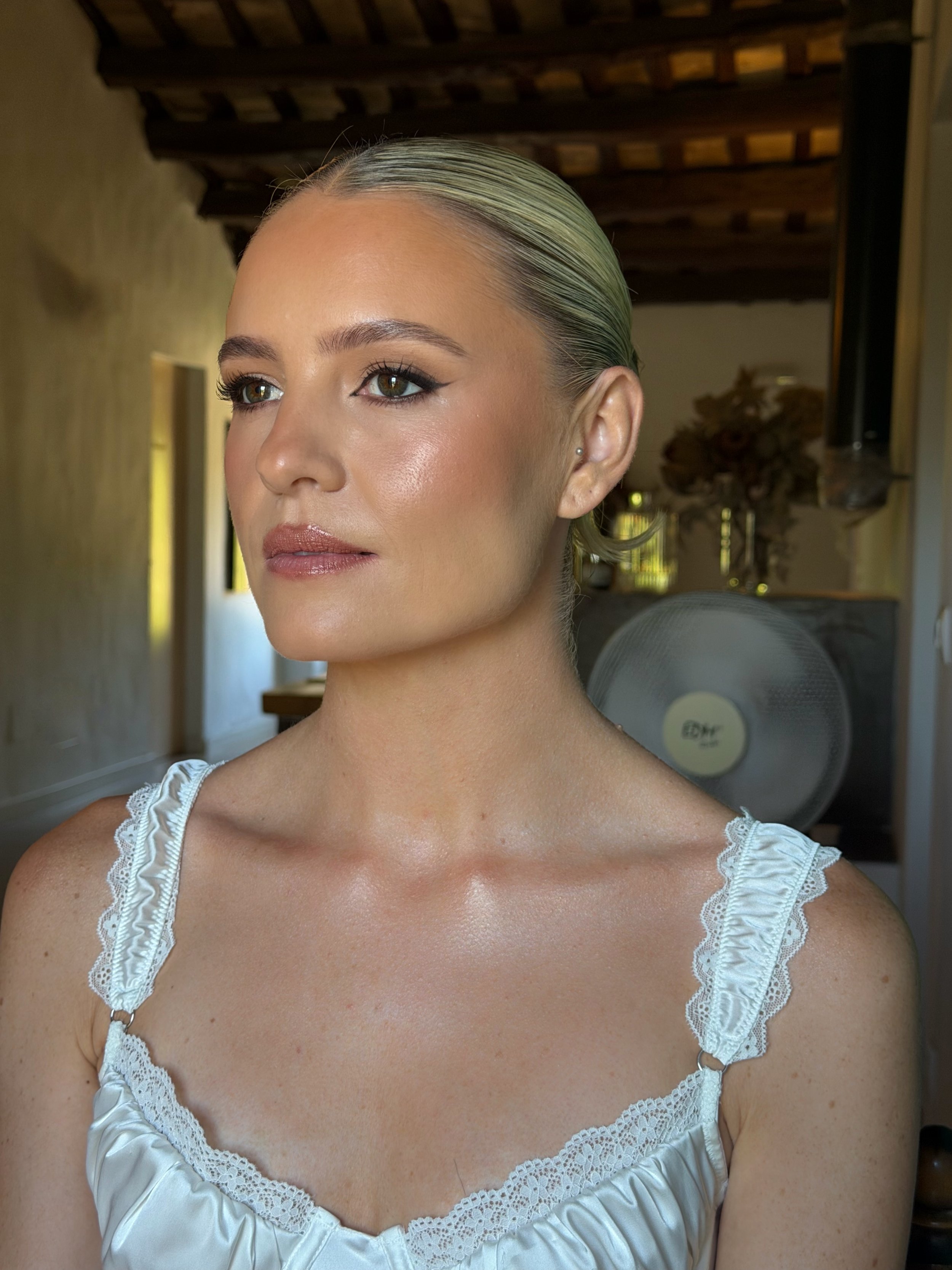 A woman with blond hair slicked back, wearing a white satin top with lace trim, looking to the side. The background shows a wooden ceiling and a fan.