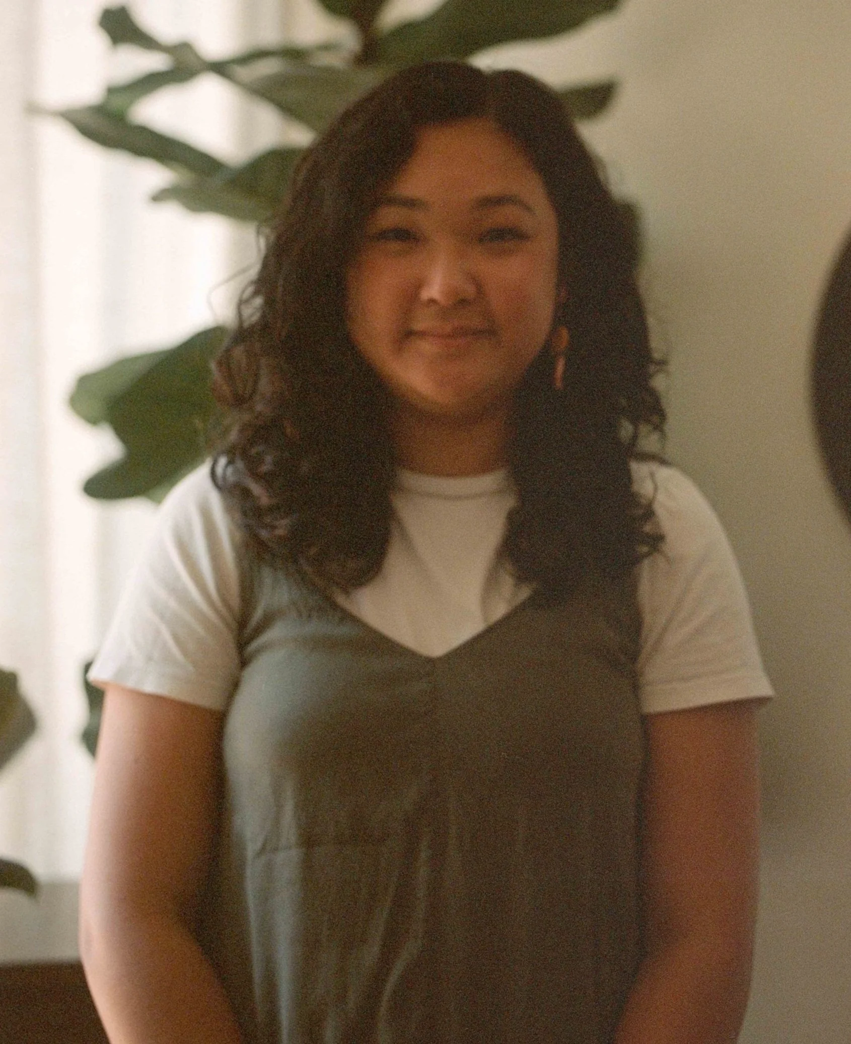 A woman with dark wavy hair smiling, wearing a white and black top, with a leafy green plant in the background.