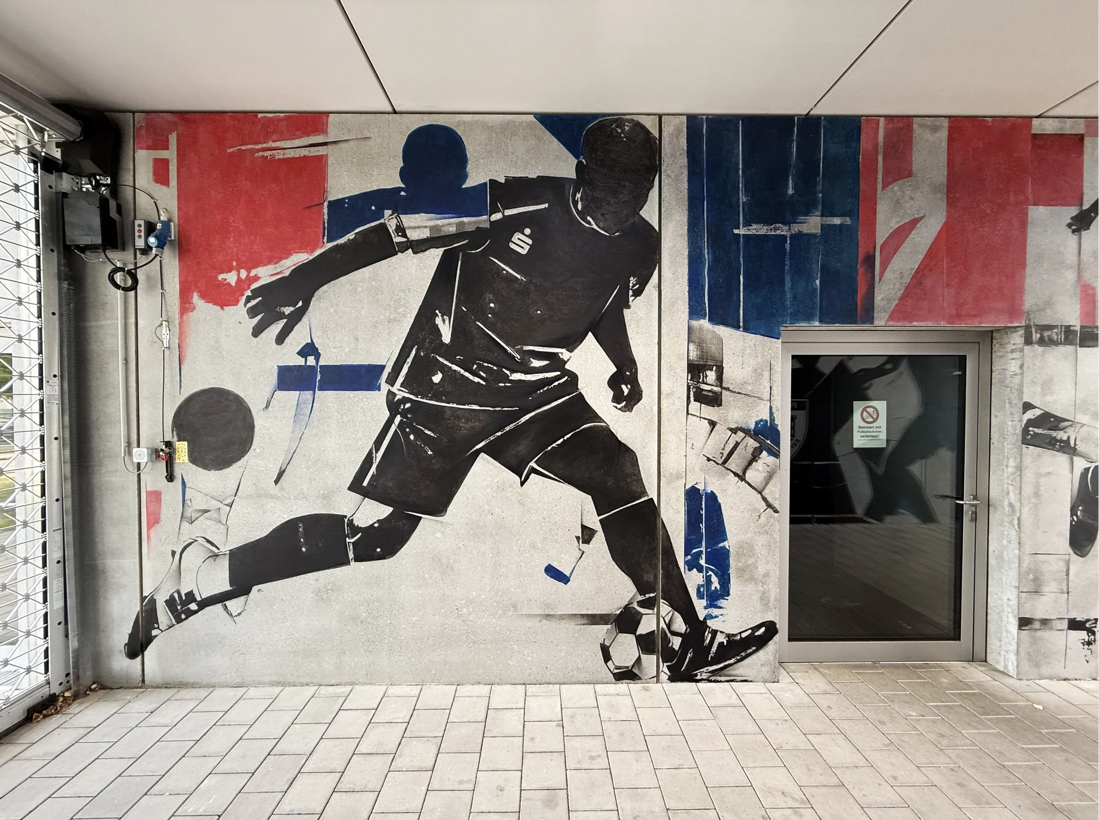 Wandmalerei eines Fußballspielers in Schwarz-Weiß, der einen Ball tritt, vor einem Hintergrund in Rot, Weiß und Blau.