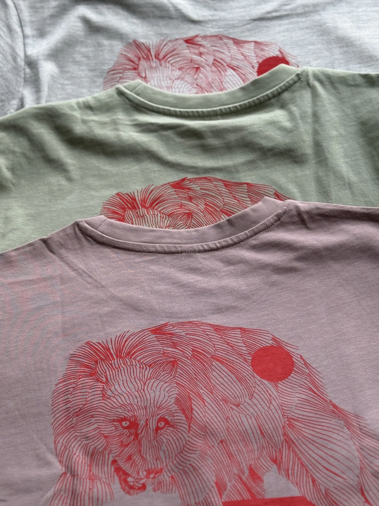 Drei übereinandergelegte T-Shirts in Grau-, Grün- und Violetttönen mit rotem Löwenmotiv.