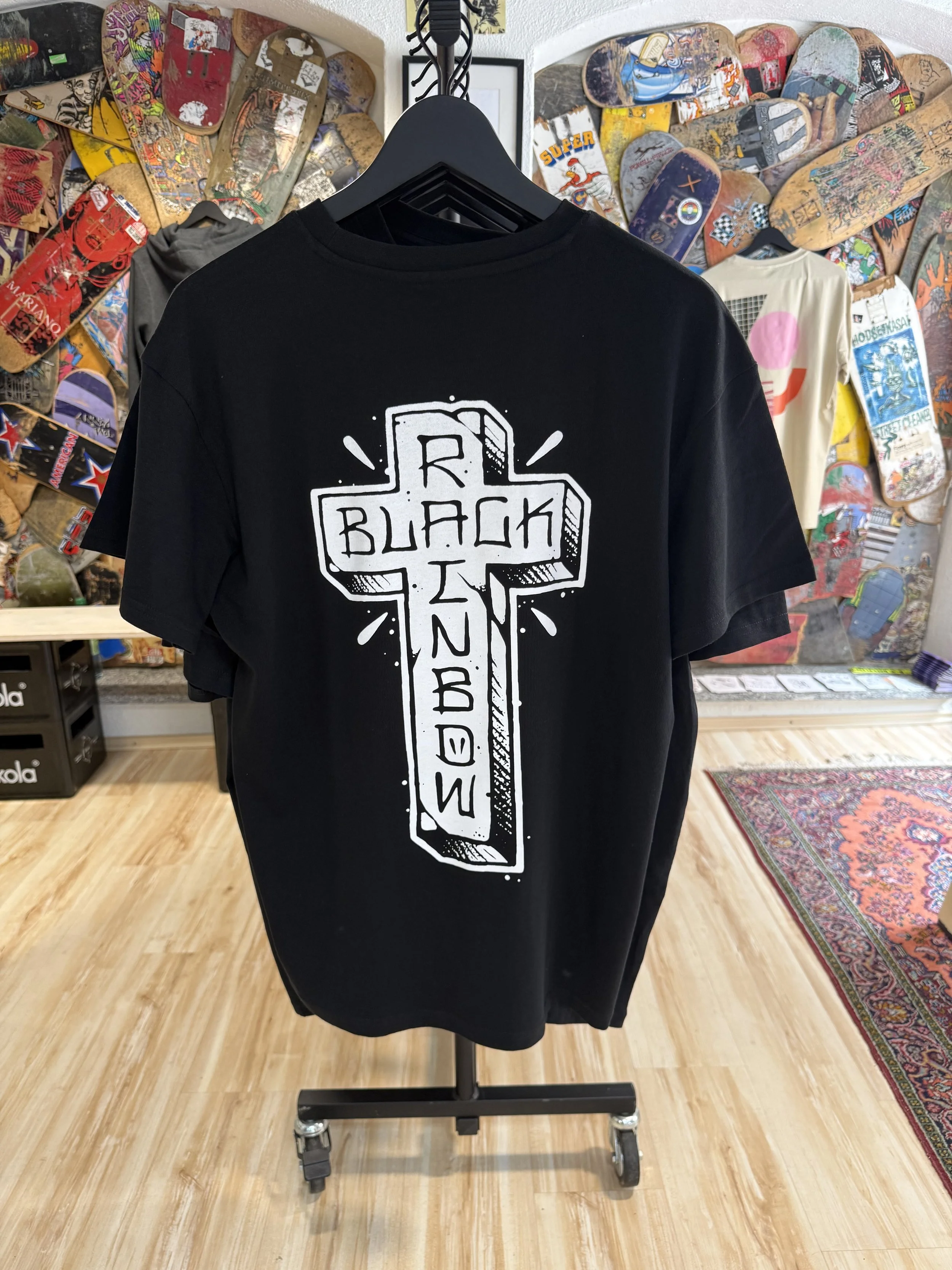 Schwarzes T-Shirt mit weißem Grafikdruck auf einem Kleiderbügel vor einer Wand voller Skateboards