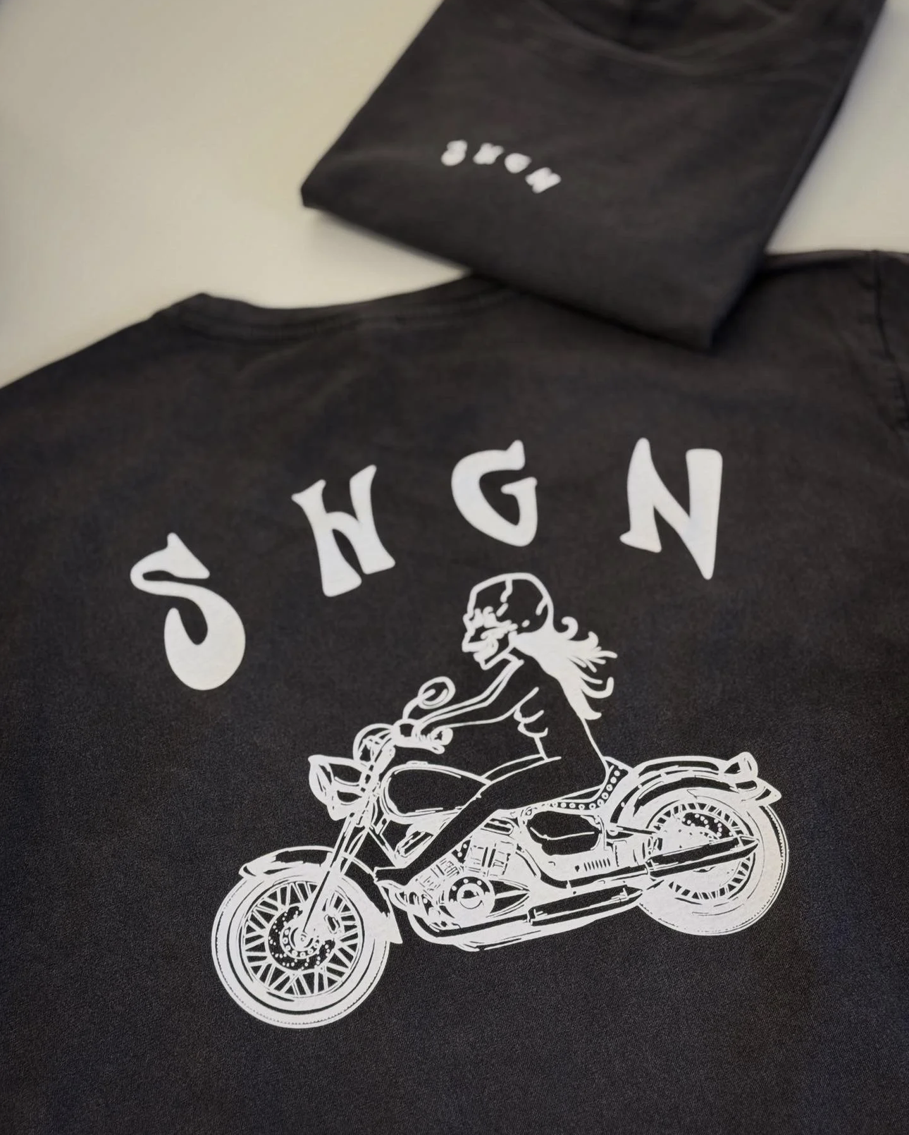 Schwarzes T-Shirt mit weißem Aufdruck „SHGN“ und einer Motorradfahrerin auf einem Cruiser-Motorrad