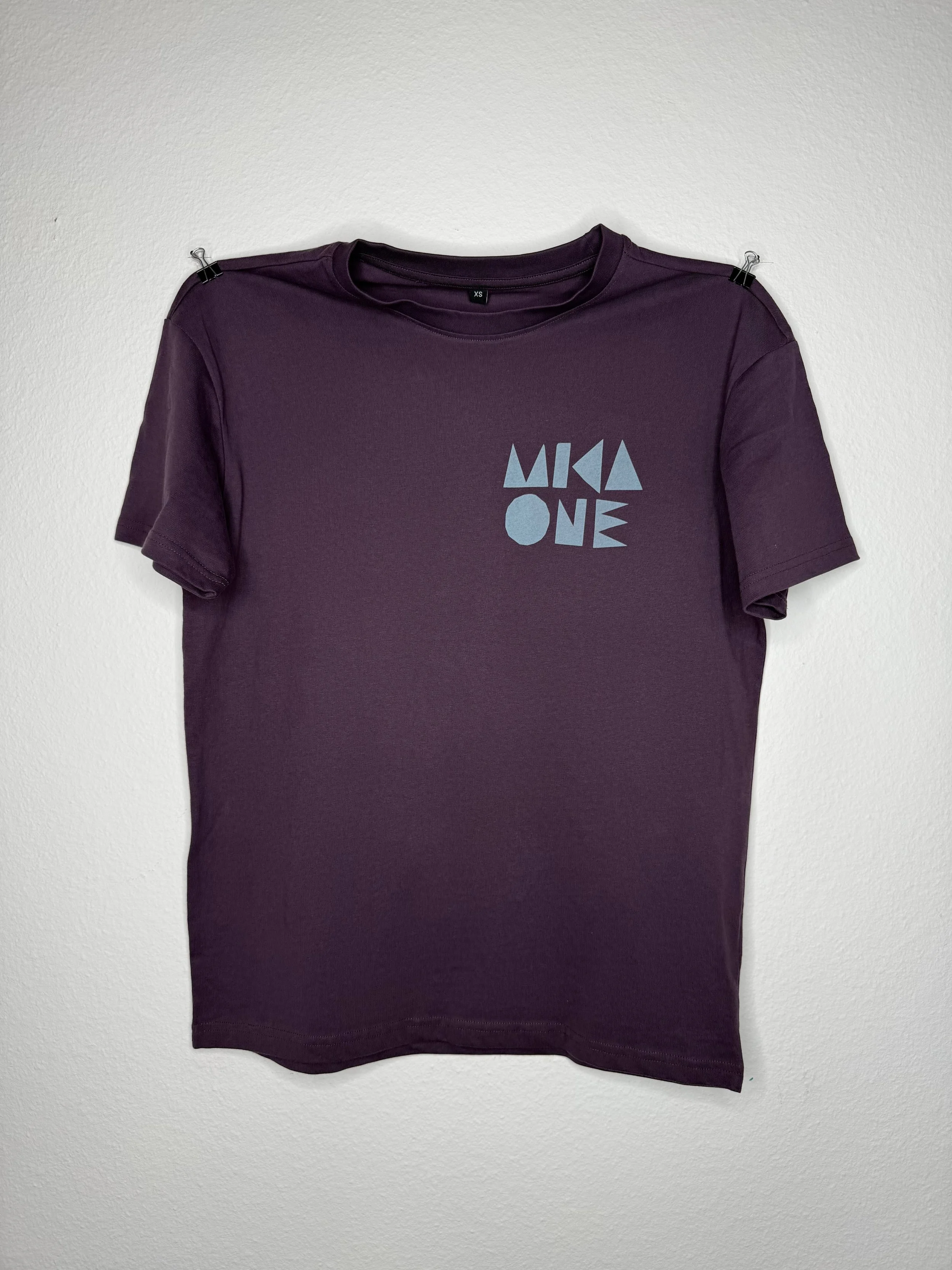 Violett-braunes T-Shirt mit Rundhalsausschnitt, kurzen Ärmeln, blauem Grafikaufdruck und Größe XS, an einer weißen Wand befestigt.