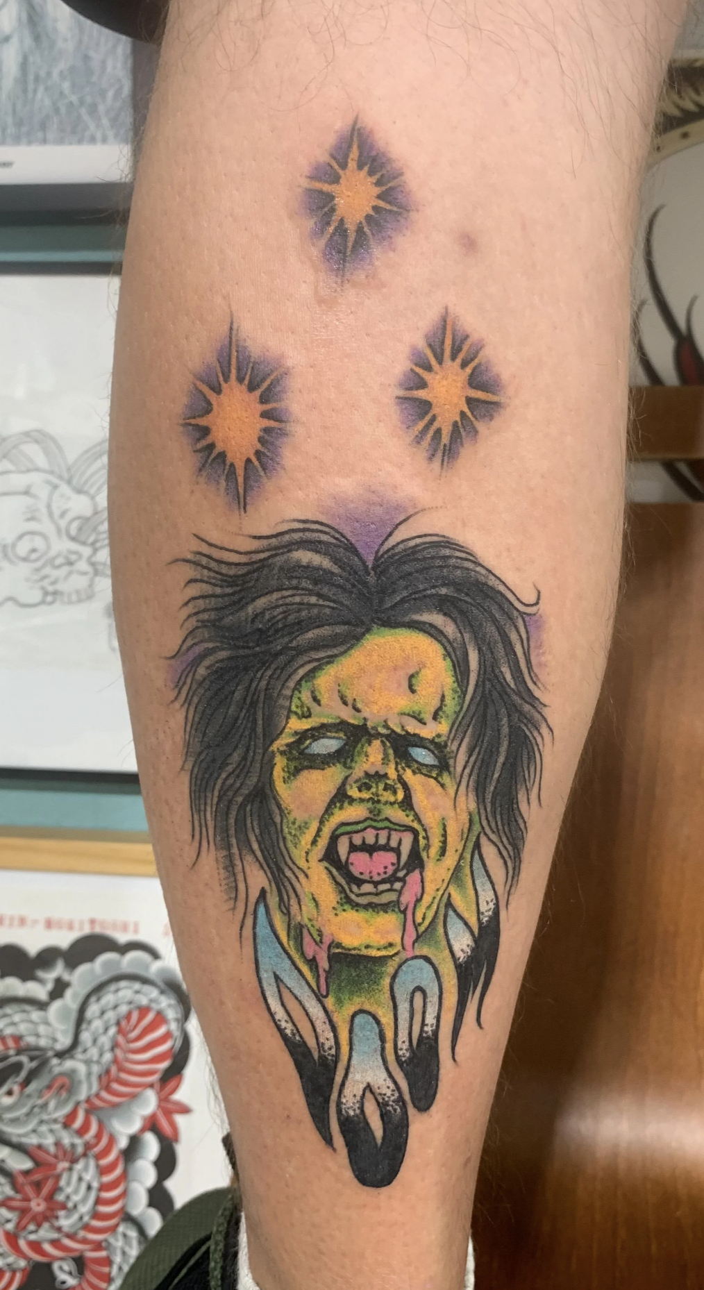 Farbiges Tattoo mit einem  Vampir- oder Monsterkopf und drei violett-goldenen Sternmotiven am Bein