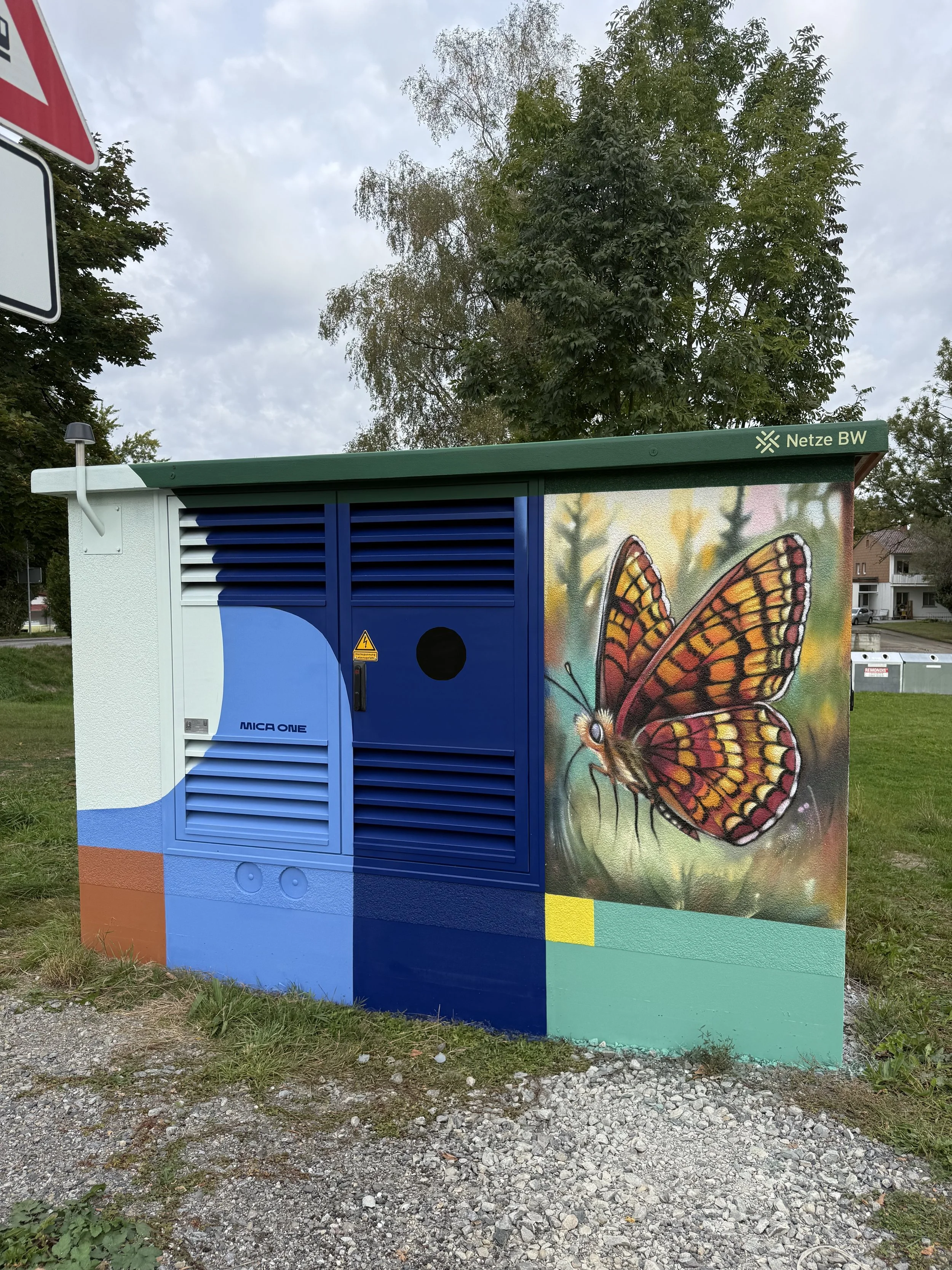 Kunstvoll bemalter Stromkasten mit einem riesigen Schmetterling auf der rechten Seite, umgeben von bunten Farbblöcken und einem Baum im Hintergrund.