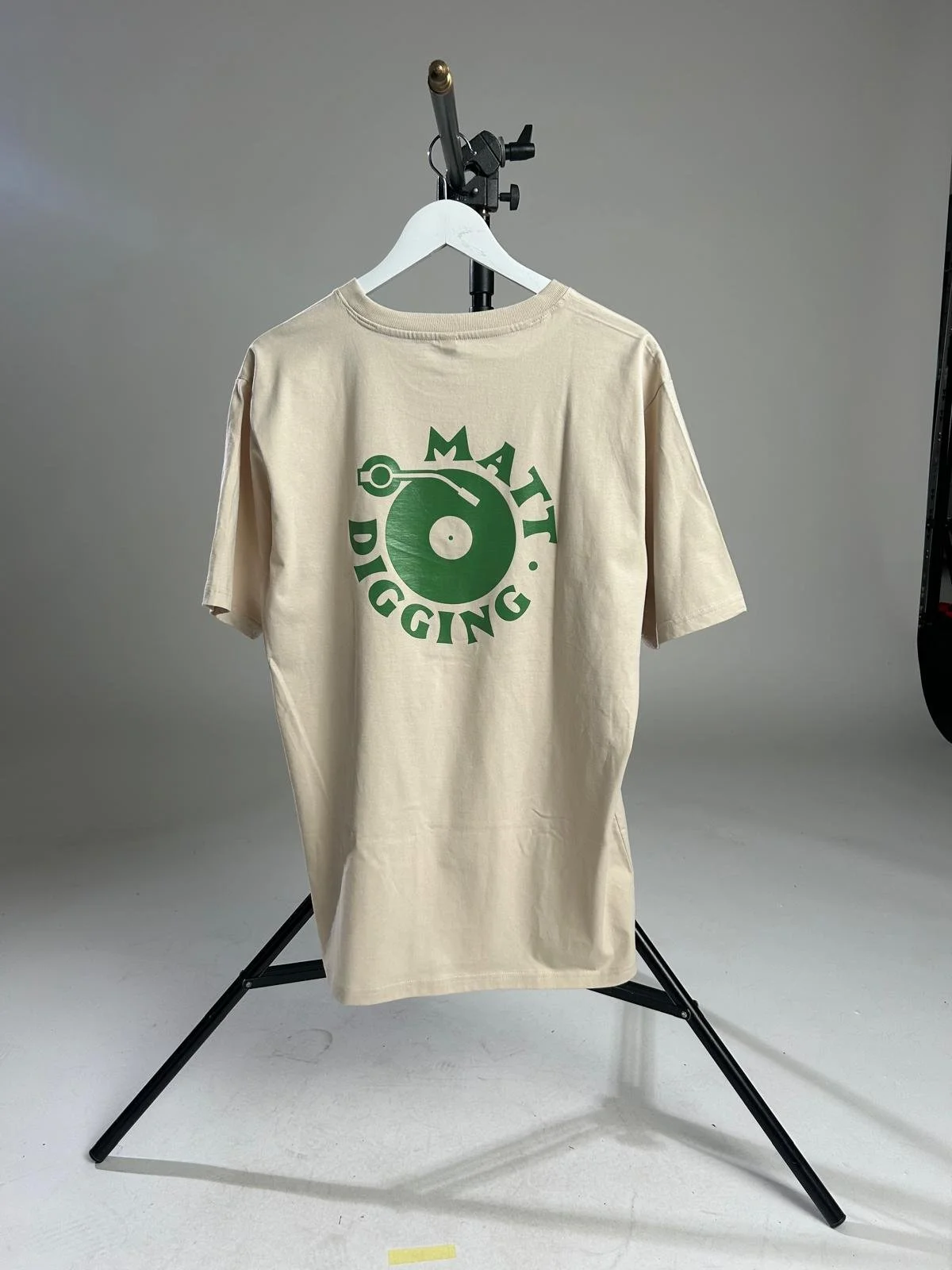 Beigefarbenes T-Shirt mit grünem Schallplatten- und „Matt Digging“-Aufdruck auf einem weißen Kleiderbügel im Fotostudio.