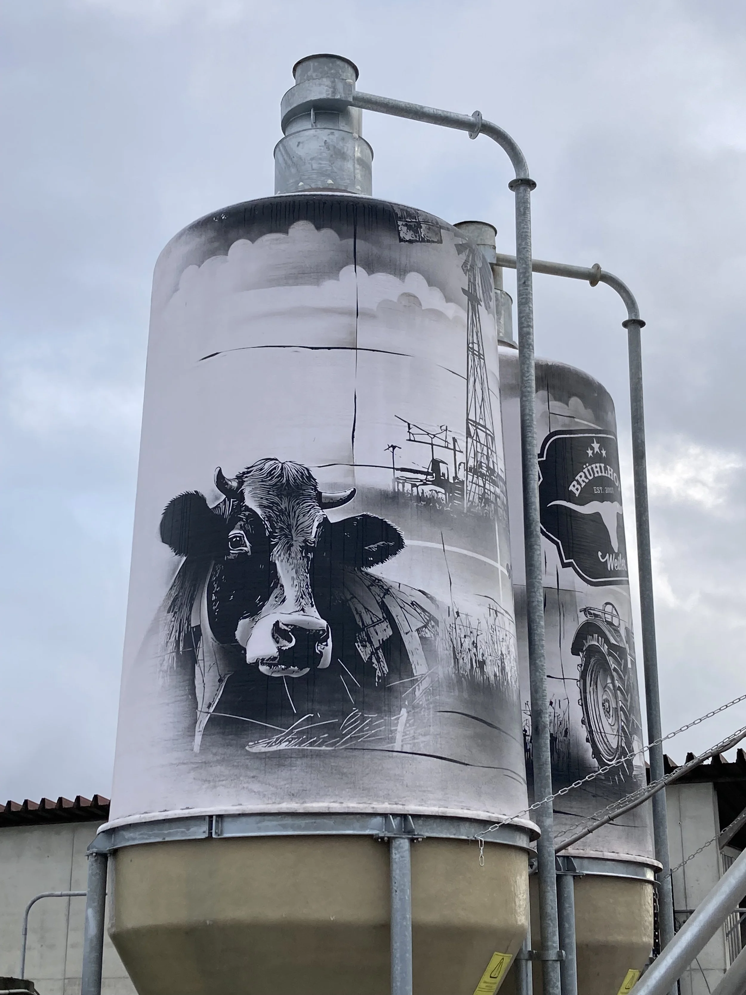 Großes Graffiti an einem Silo zeigt eine Zeichnung eines Kuhkopfes, im Hintergrund ist ein Bauernhof mit Windmühle und landwirtschaftlicher Geräte. 