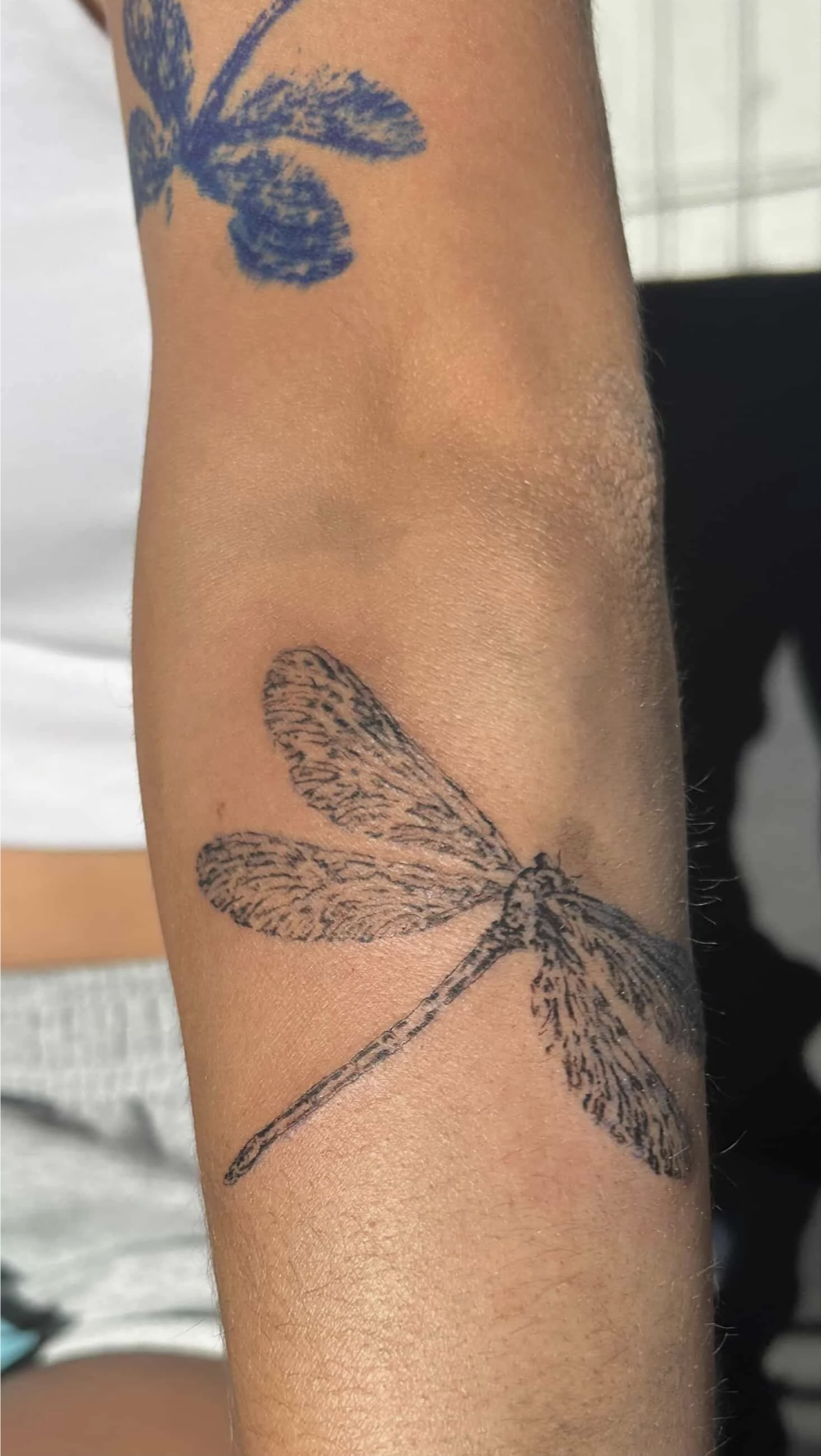 Nahaufnahme eines Unterarms mit einem Libellen-Tattoo und einem teilweise sichtbaren blauen Blumentattoo.