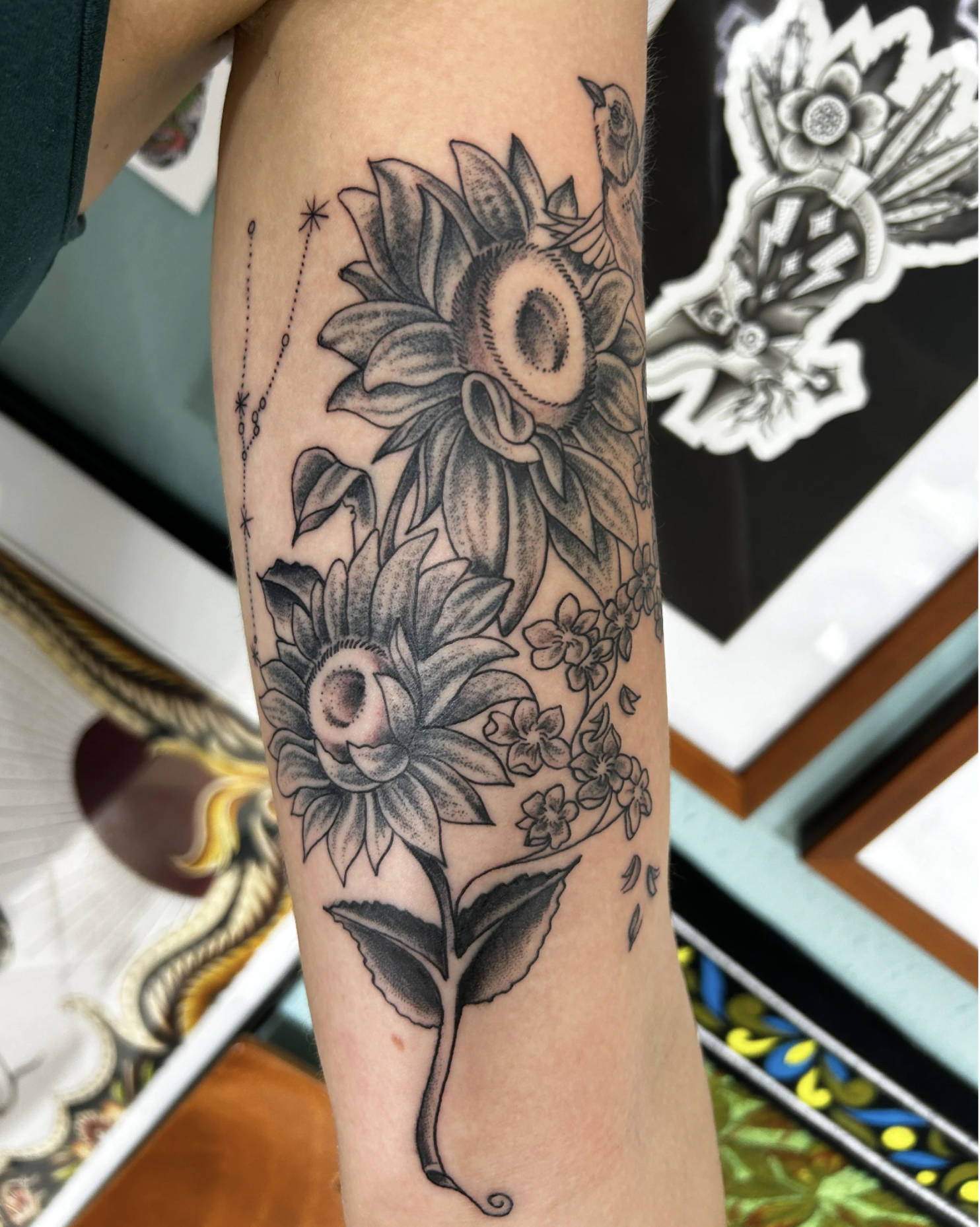 Schwarz-graues Blumen-Tattoo mit großen Sonnenblumen, kleinen Blüten, Blättern und dekorativen Linien auf einem Unterarm