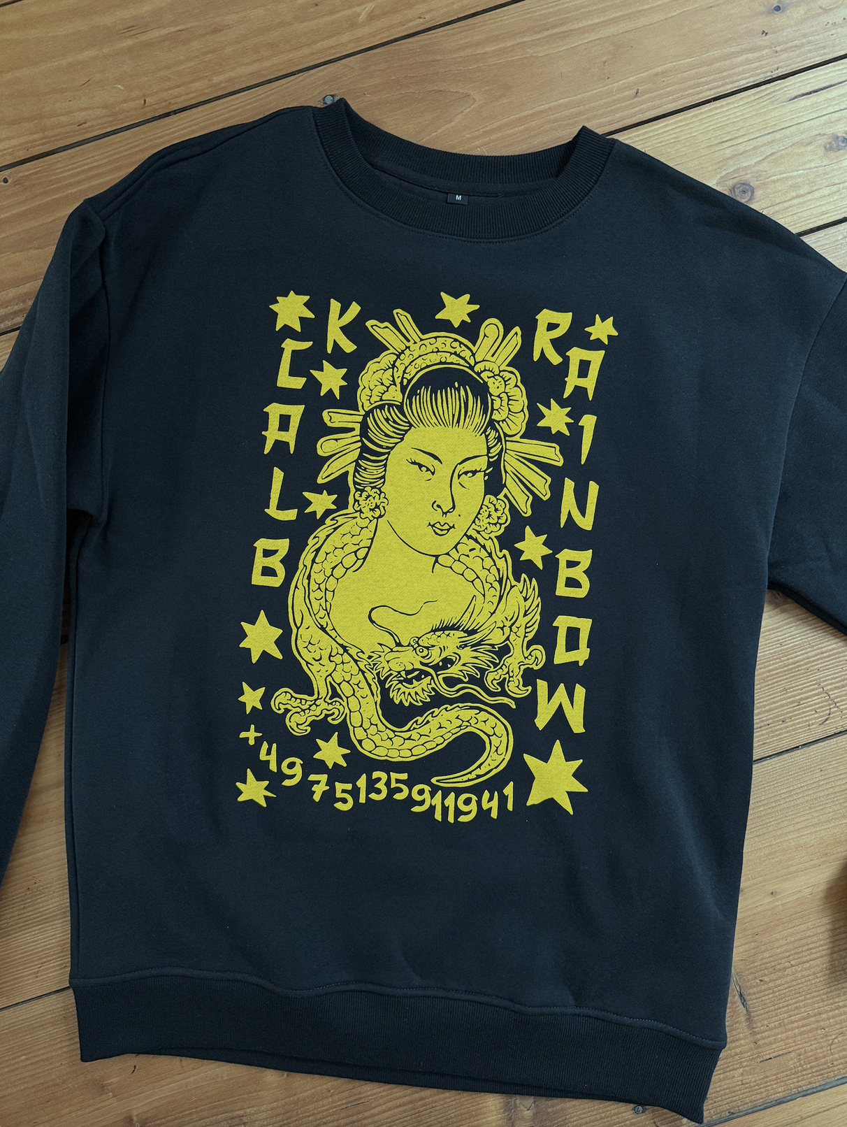 Schwarzes Sweatshirt mit gelbem Motiv einer japanischen Geisha, eines Drachens, Sternen und asiatischen Schriftzeichen auf Holzboden.