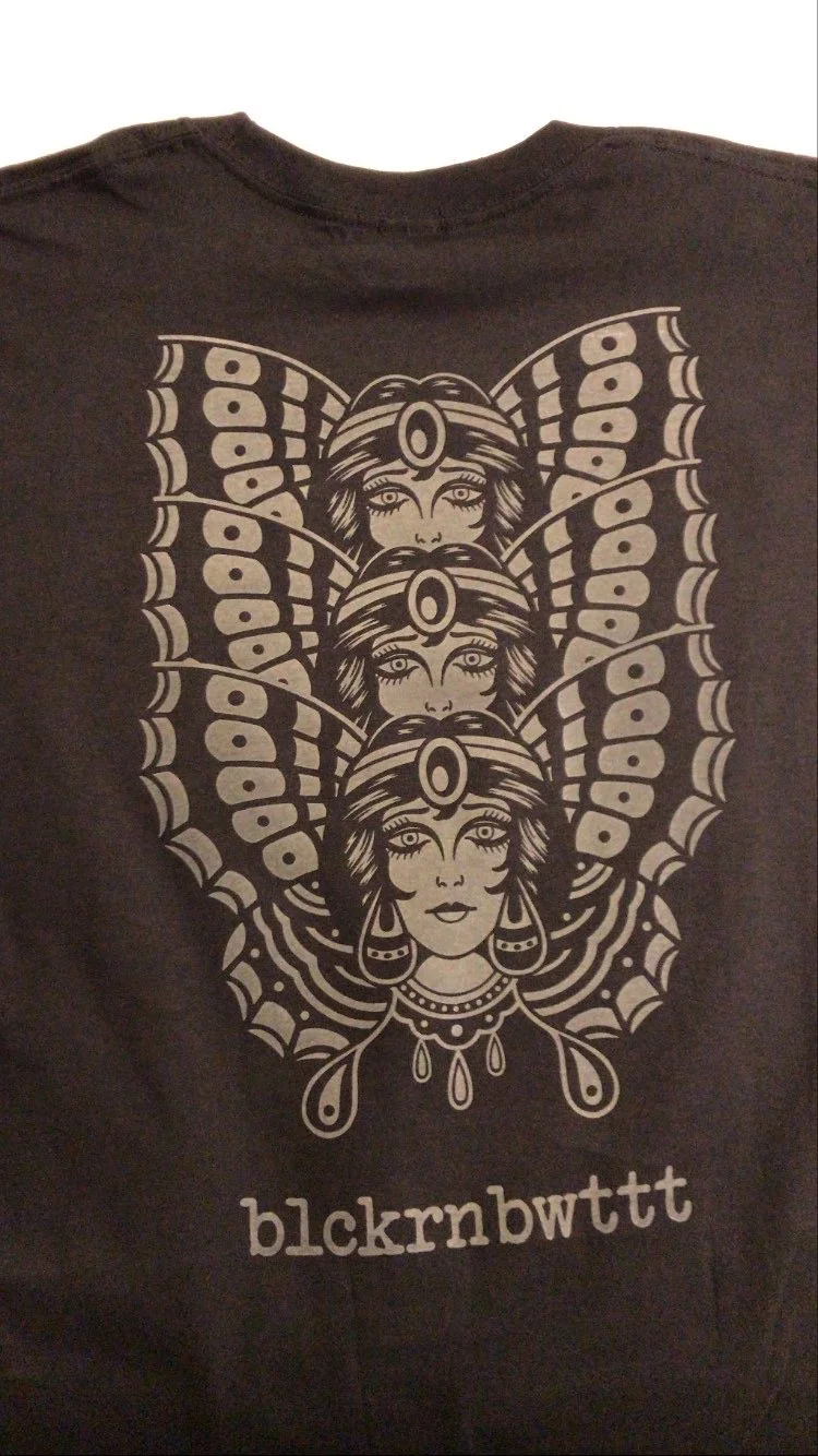 Schwarzes T-Shirt mit grafischem Motiv aus drei stilisierten Frauenporträts mit Flügeln und dem Schriftzug „blckrnbwttt“.