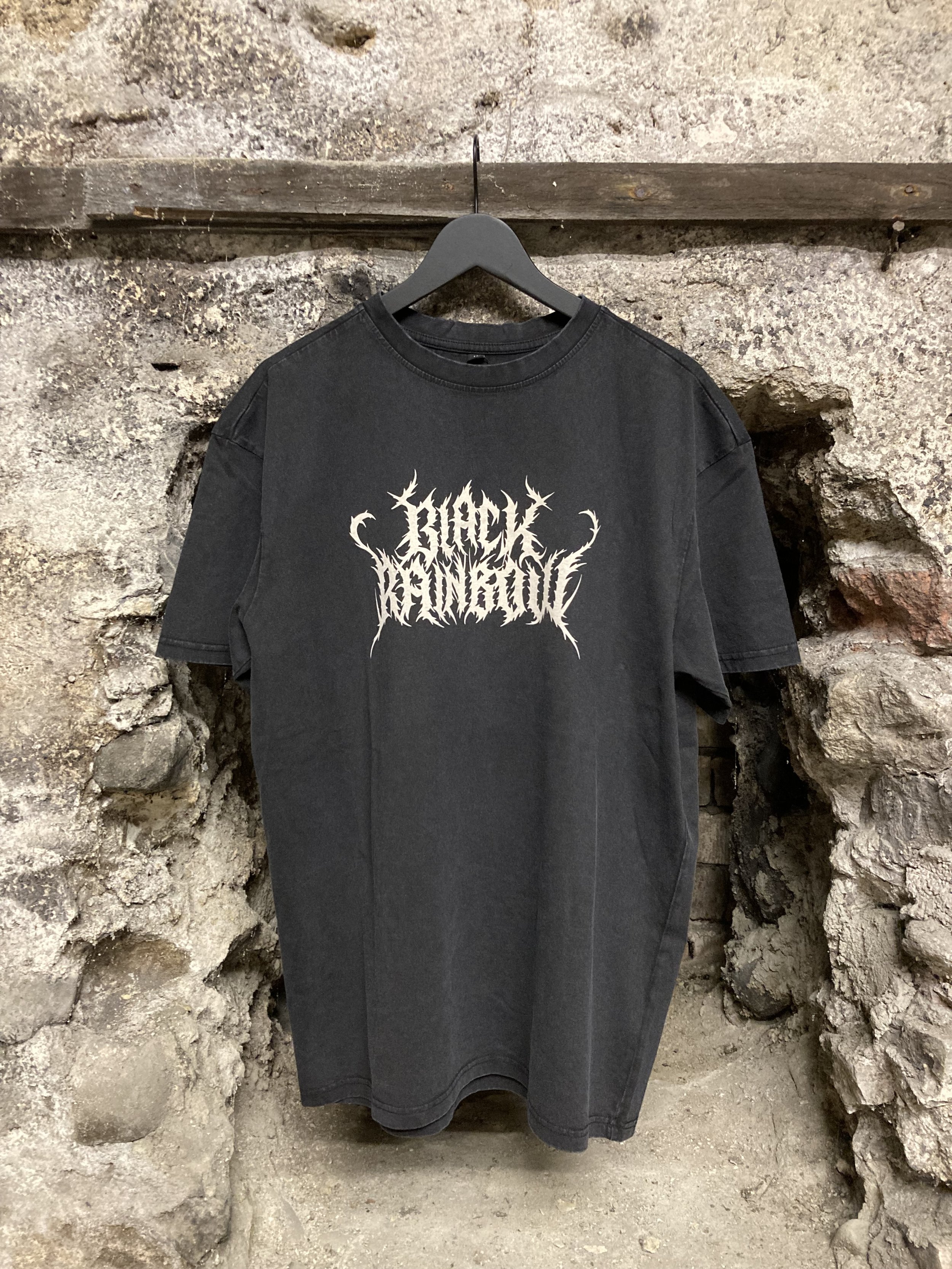 Schwarzes T-Shirt mit weißem Death-Metal-Schriftzug und Dornenmuster, an einem Kleiderbügel vor einer rustikalen Steinmauer.