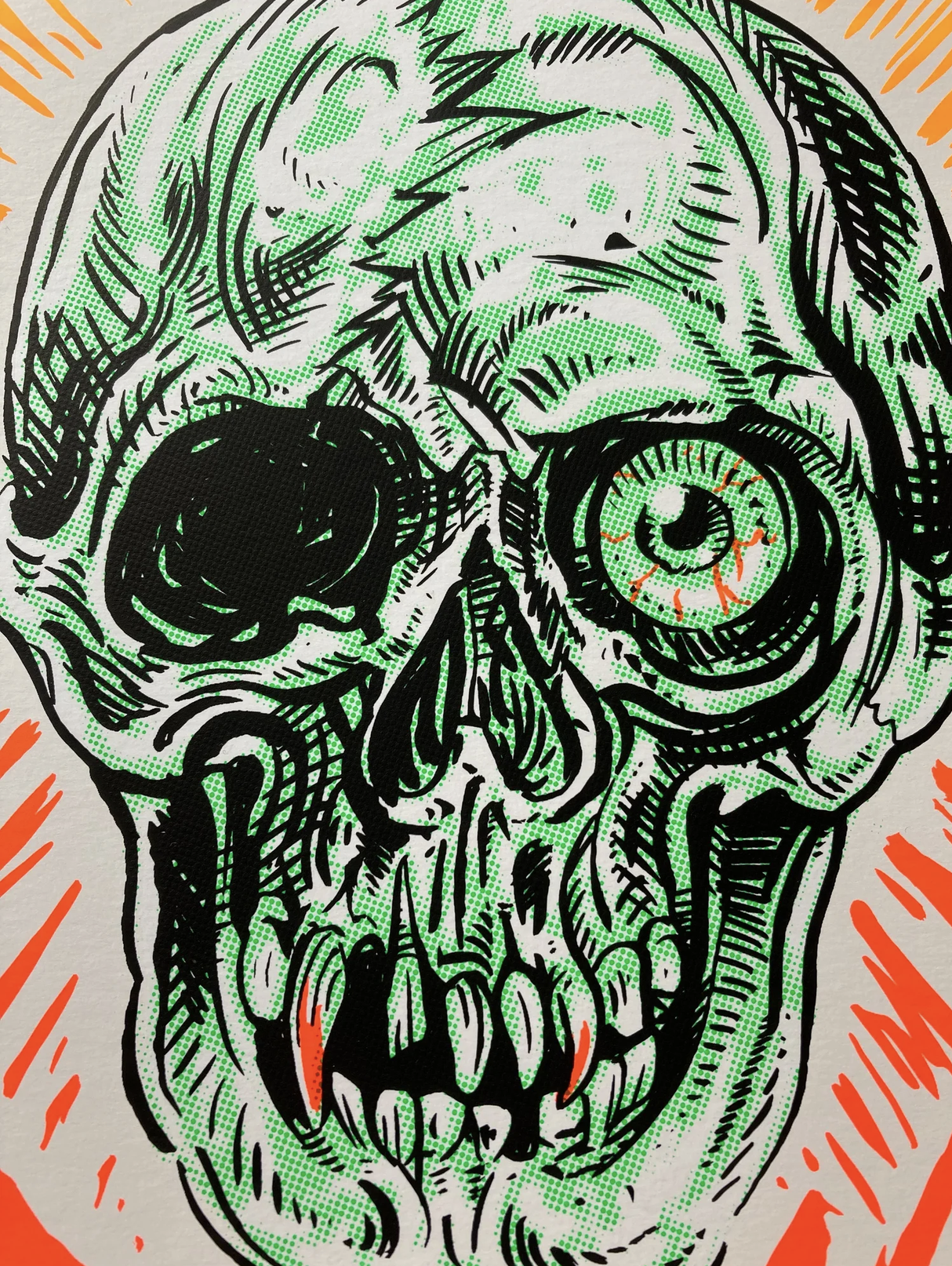 Nahaufnahme eines Siebdrucks eines grimmigen Totenkopfes mit einem großen grünen Auge, schwarzen Linien und orangefarbenen Akzenten.