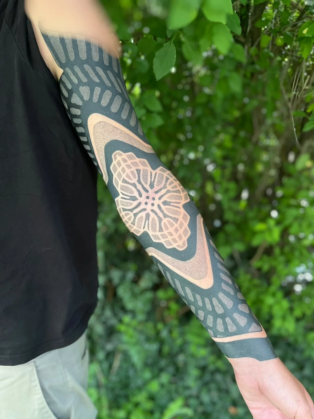 Nahaufnahme eines Arms mit großflächigem geometrischem Tattoo und floralem Mandala-Motiv vor grünem Laub.