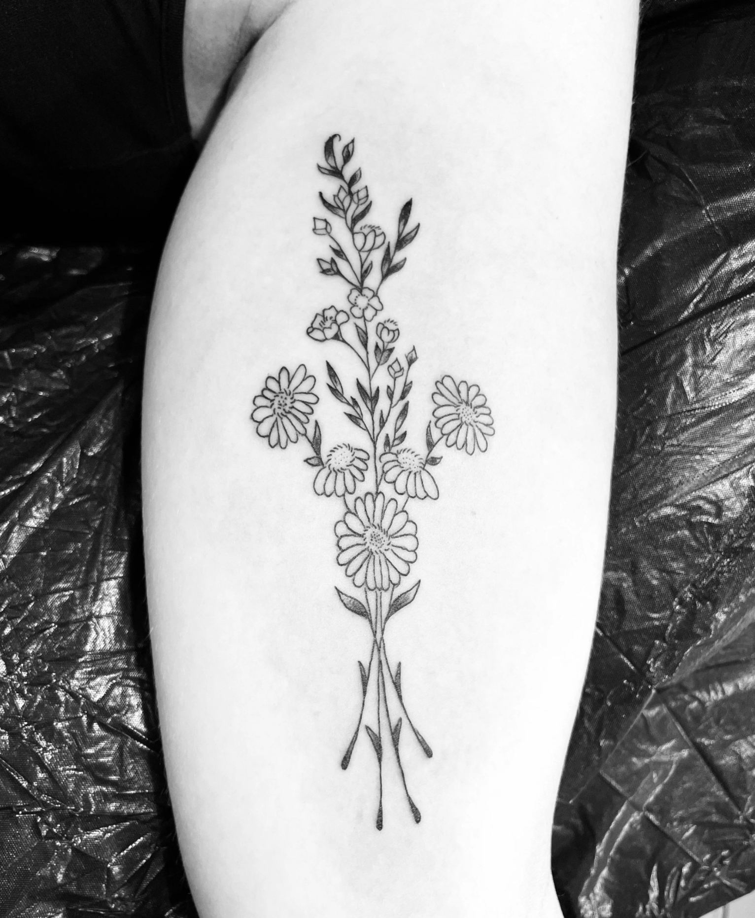 Schwarz-weißes florales Tattoo mit Gänseblümchen, Blüten und Blättern auf einem Unterarm.