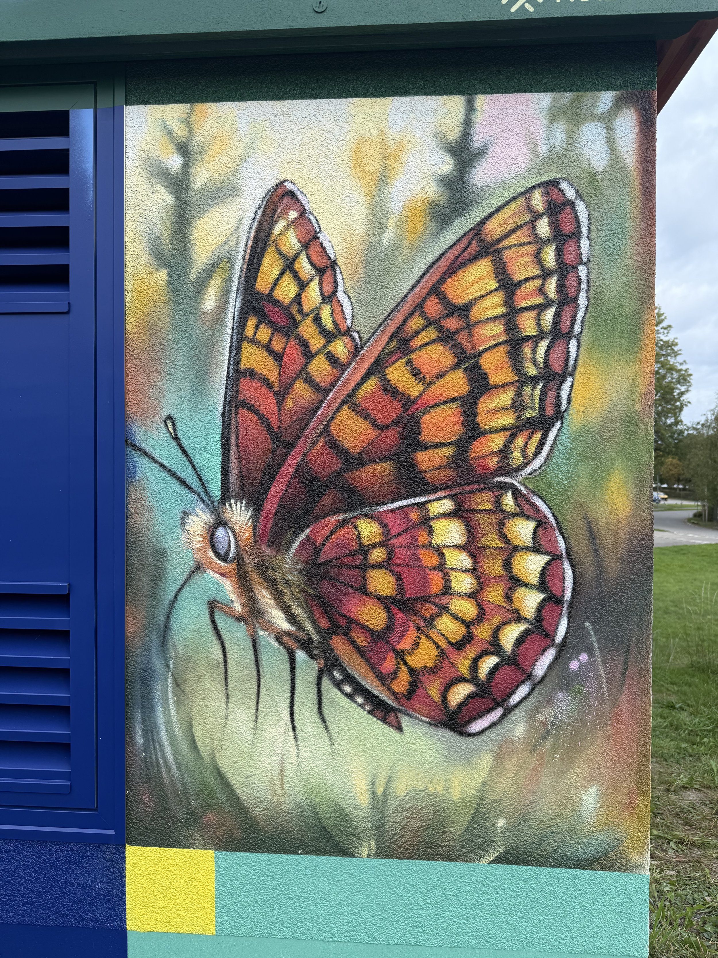 Das Bild zeigt eine Wandmalerei einer stilisierten Schmetterlingsflügel in orange, gelb und schwarz, auf einer grauen Wand mit bokeh-ähnlichem Hintergrund und einigen Bäumen im Hintergrund.
