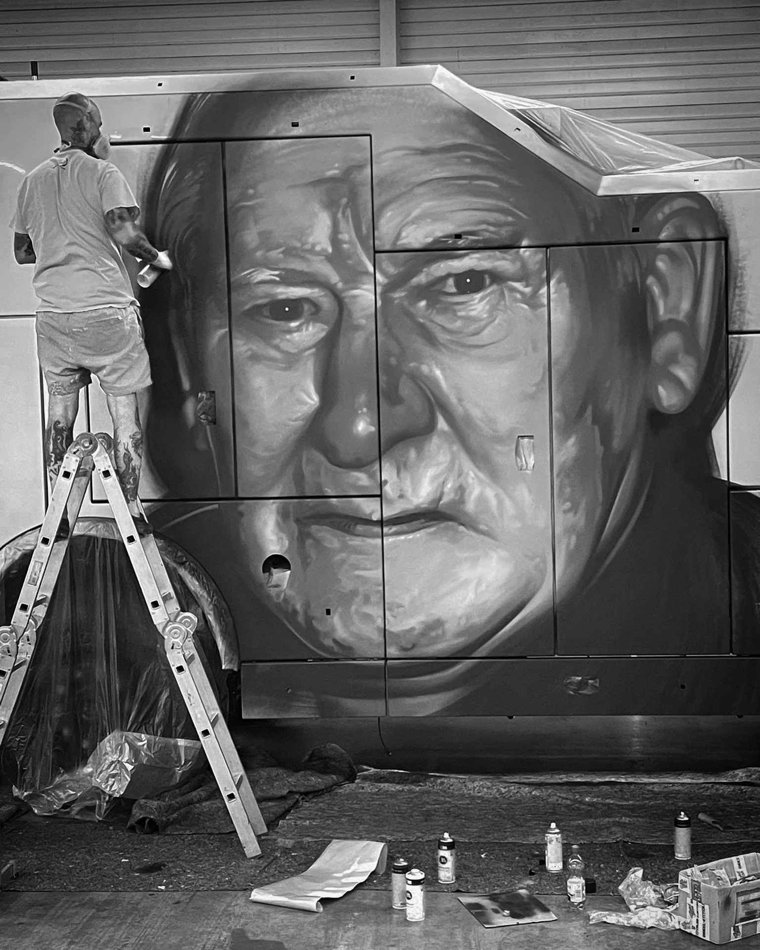 Ein Mann arbeitet an einem Graffiti-Bild eines menschlichen Gesichts auf der Seite eines Fahrzeugs, umgeben von Malwerkzeugen und Sprühdosen auf dem Boden.
