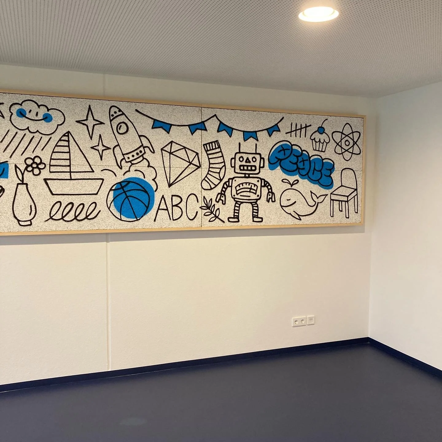 Kunstwerk mit Handzeichnungen als Kinder- und Freizeitmotive, inklusive Rakete, Ball, Schiff, Gitarre, Roboter, Delfin, Wolken, Cupcake, Atommodell und die Buchstaben ABC, auf einer weißen Wand im Raum.