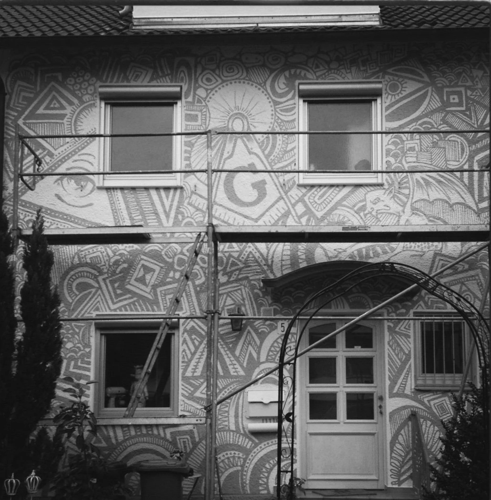 Schwarz-weißes Foto eines mit geometrischen Wandmalereien verzierten Hauses, eingerahmt von Baugerüst und Pflanzen.