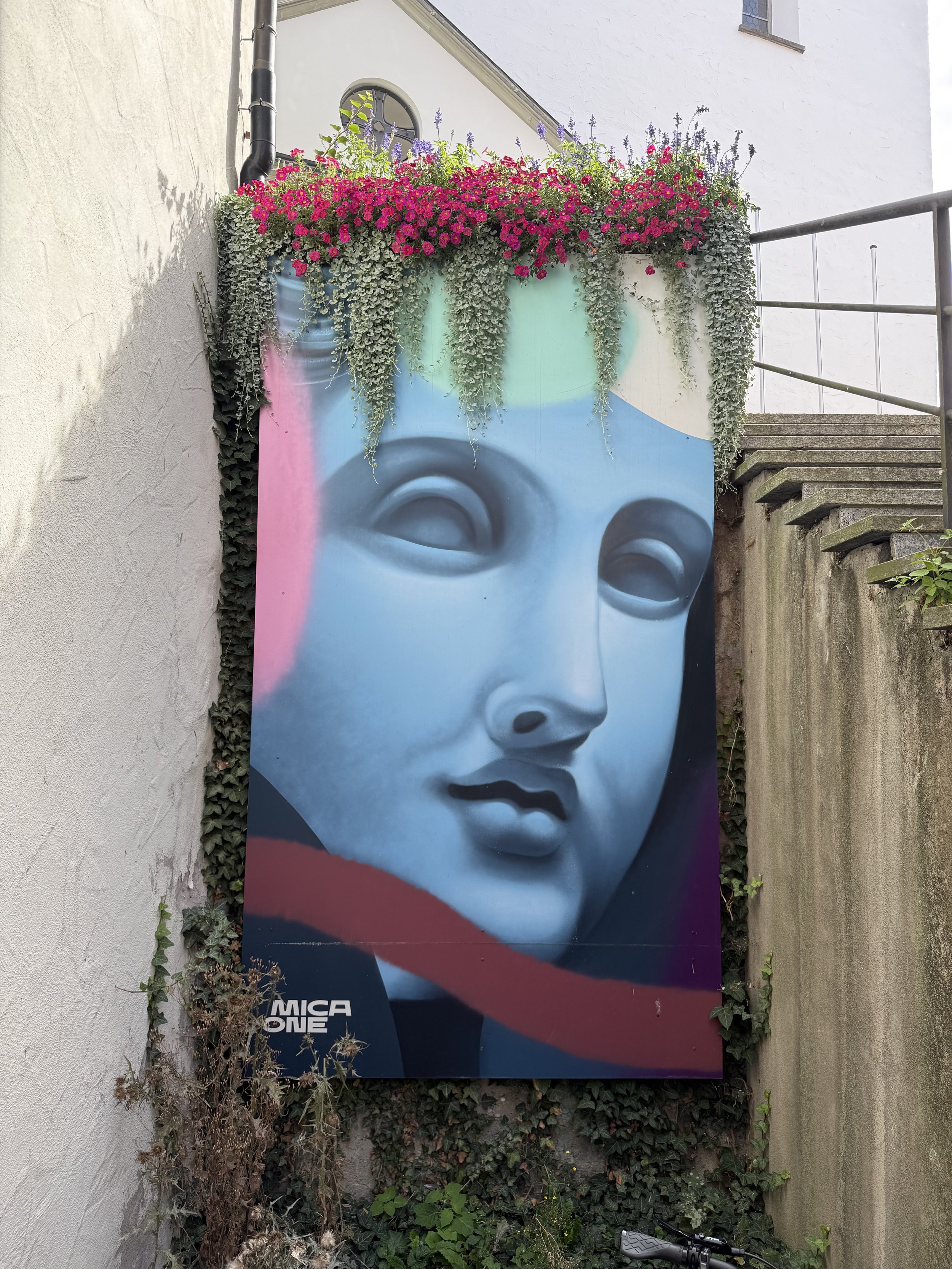 Streetartporträt einer blauen, stilisierten menschlichen Gesichtsdarstellung, umgeben von buntem Blumenarrangement und Kletterpflanzen vor einer weißen Hauswand.
